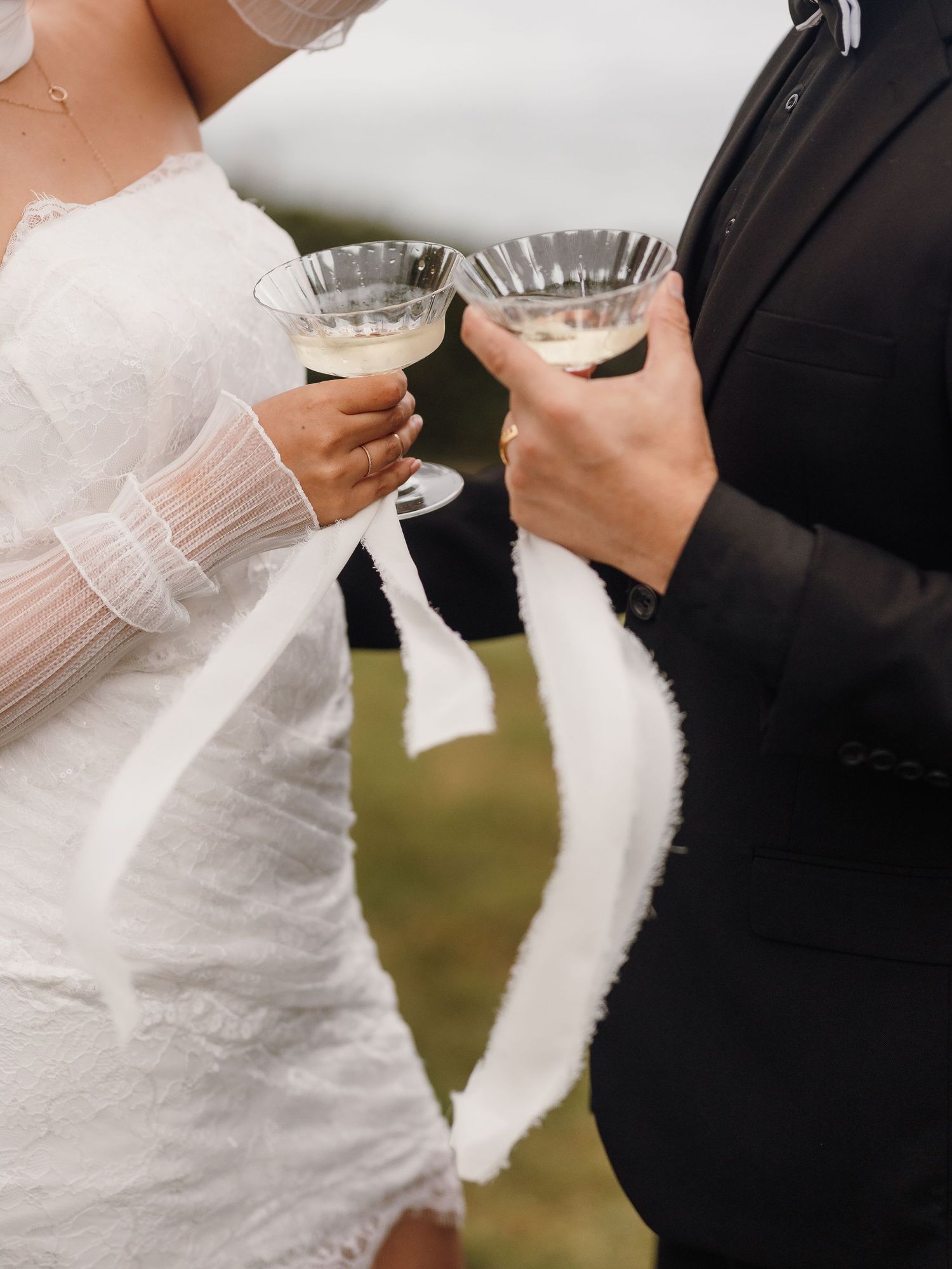 Bride and groom clinking coupe glasses in a wedding toast, wearing white lace dress and black suit outdoors.