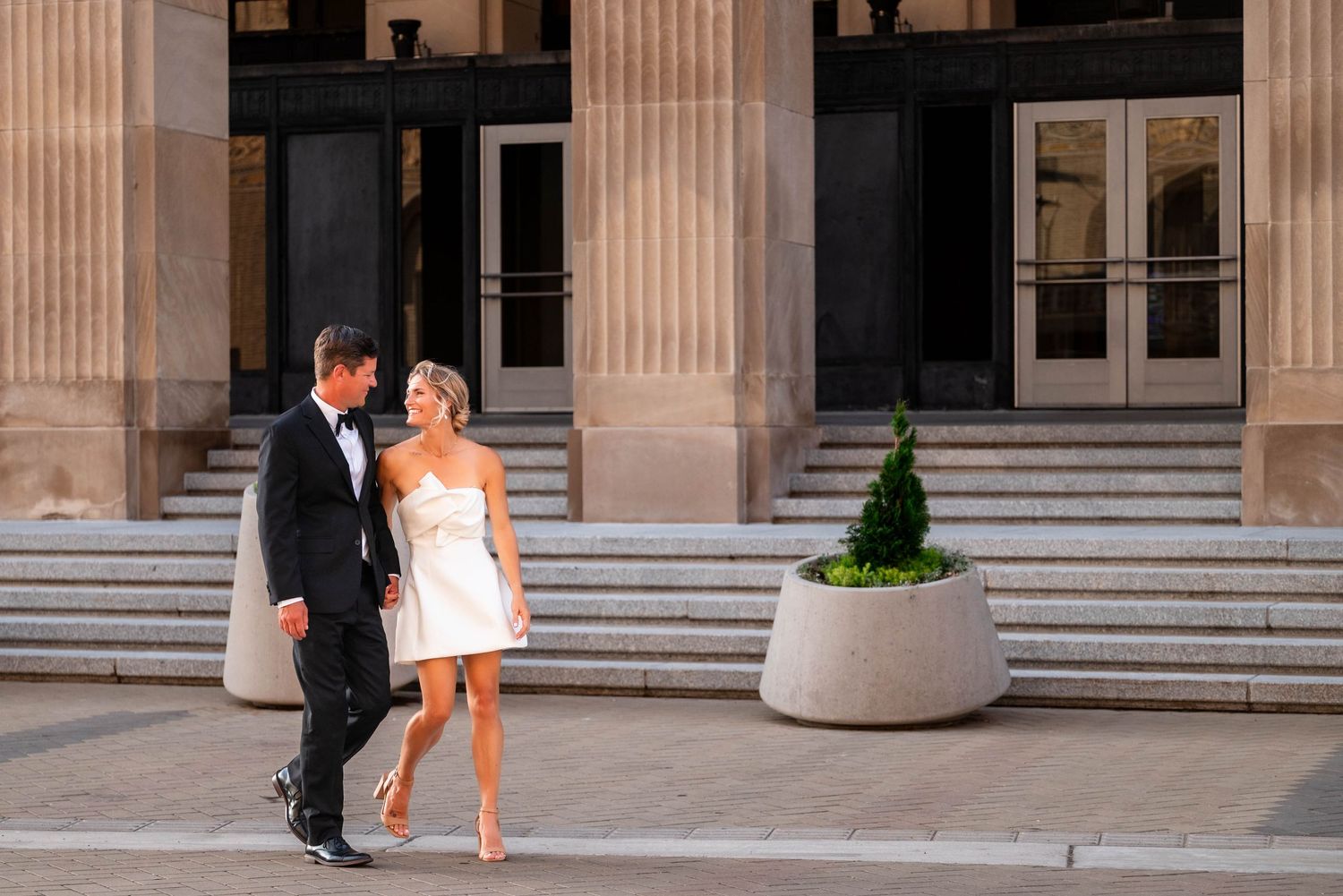Couple in wedding attire walking outside a grand building, groom in tuxedo and bride in short white dress.