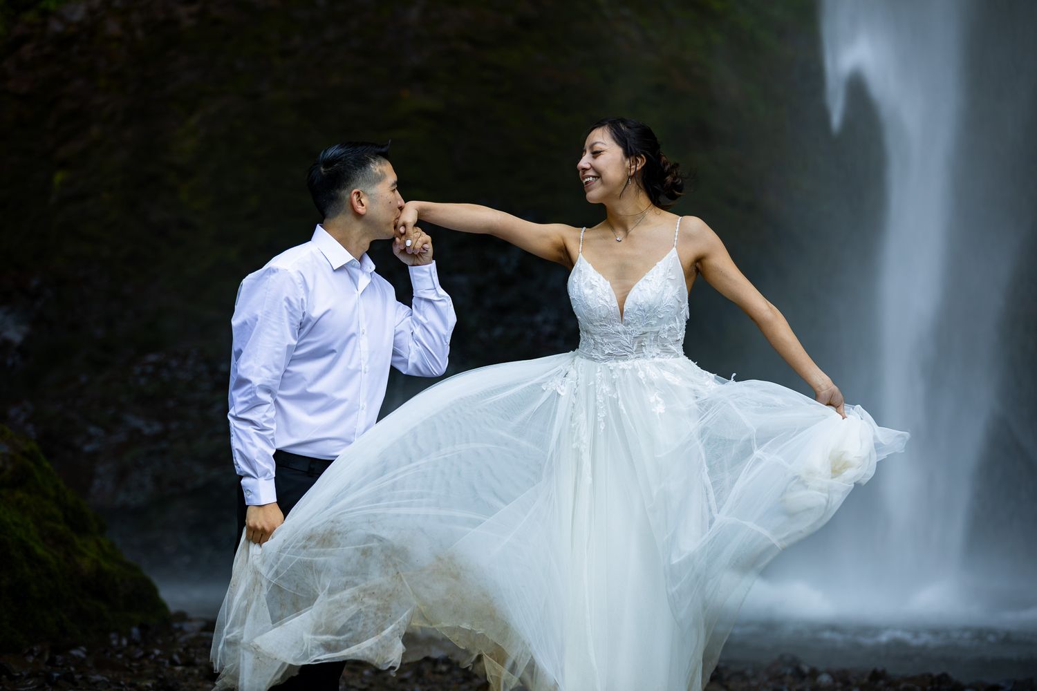 Couple in wedding attire poses romantically before an Oregon waterfall, bride in flowing white gown, groom kissing her hand.