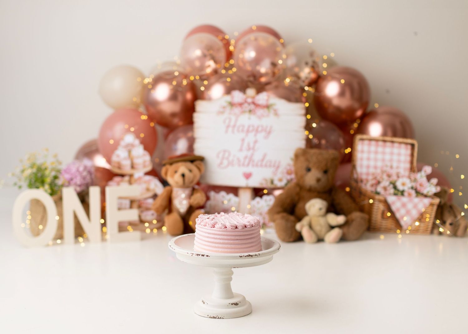 Pink first birthday setup with rose gold balloons, teddy bears, ONE sign, and a smash cake on a white pedestal.