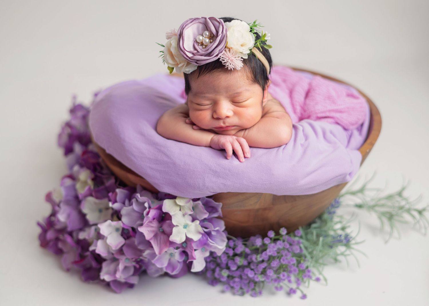 Newborn baby sleeping peacefully in a wooden bowl with purple floral wreath headband and lavender blooms.