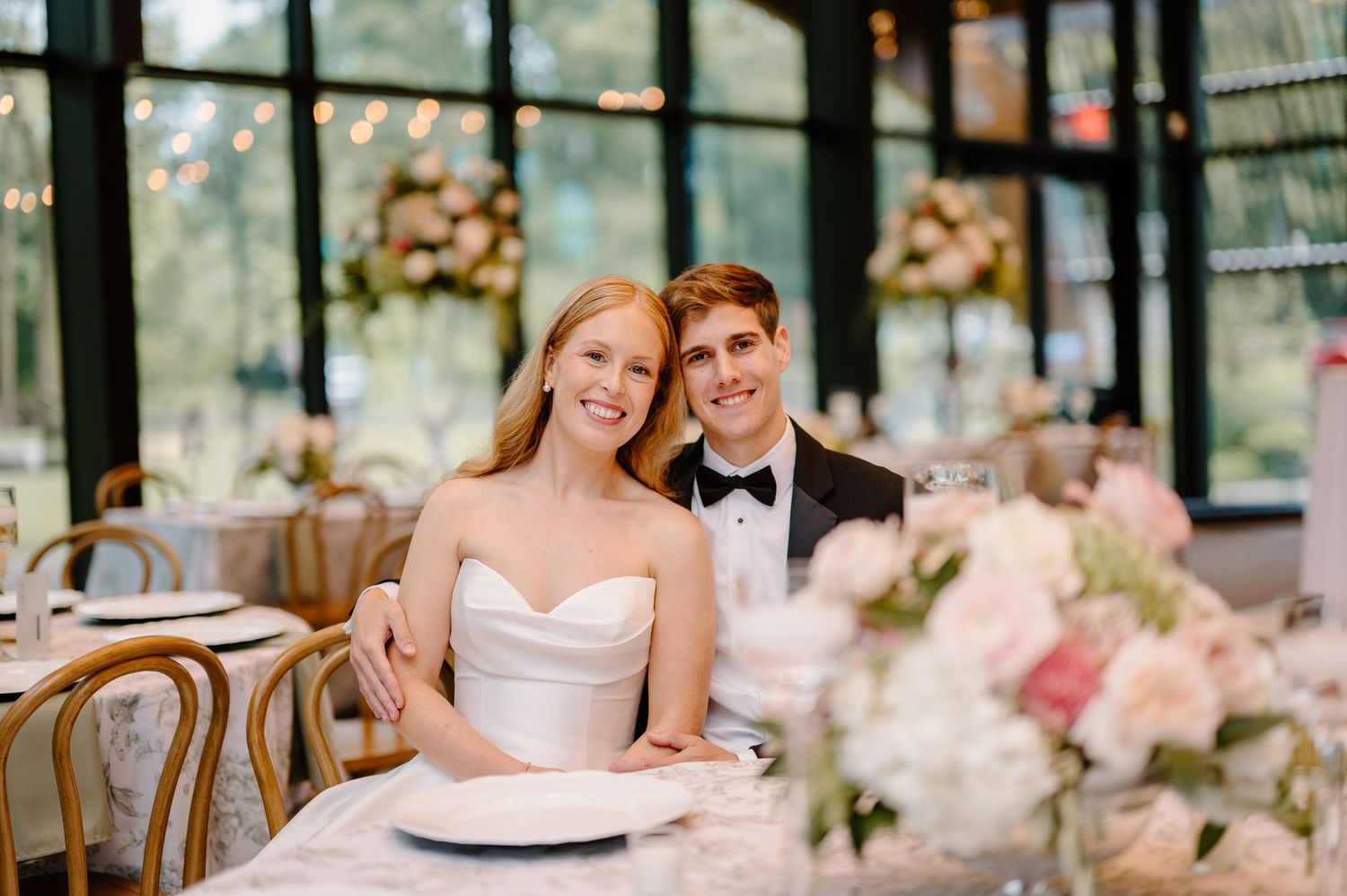 Bride and groom smiling at sweetheart table during elegant wedding reception with floral centerpieces.