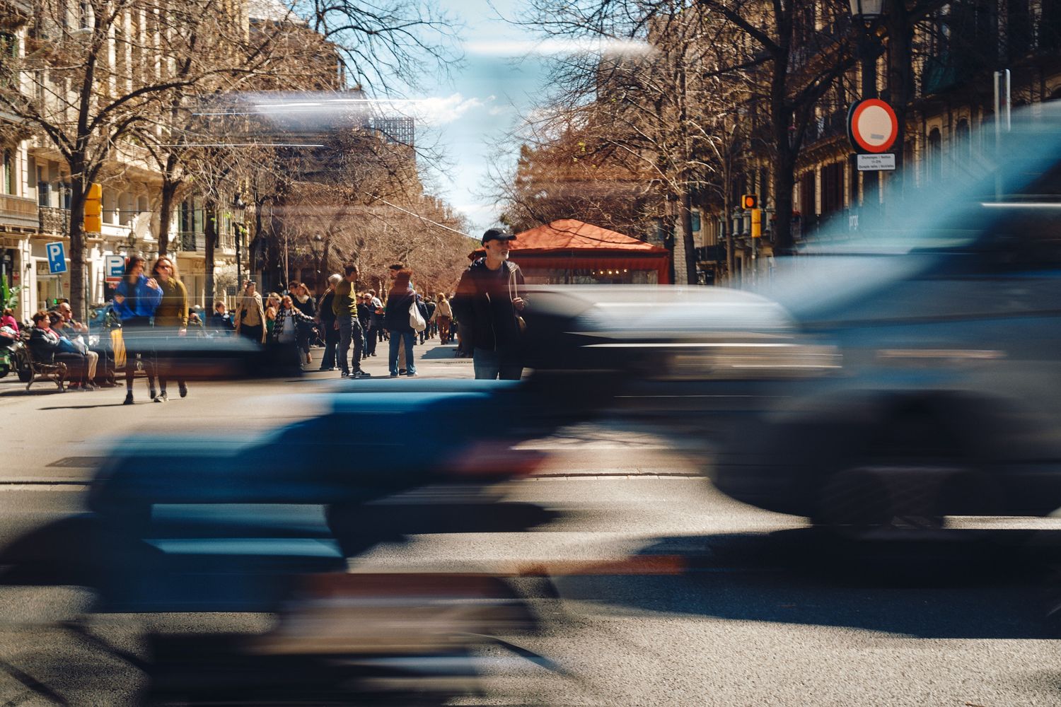 Blurred motion of vehicles and pedestrians on a busy urban street lined with trees on a sunny day.