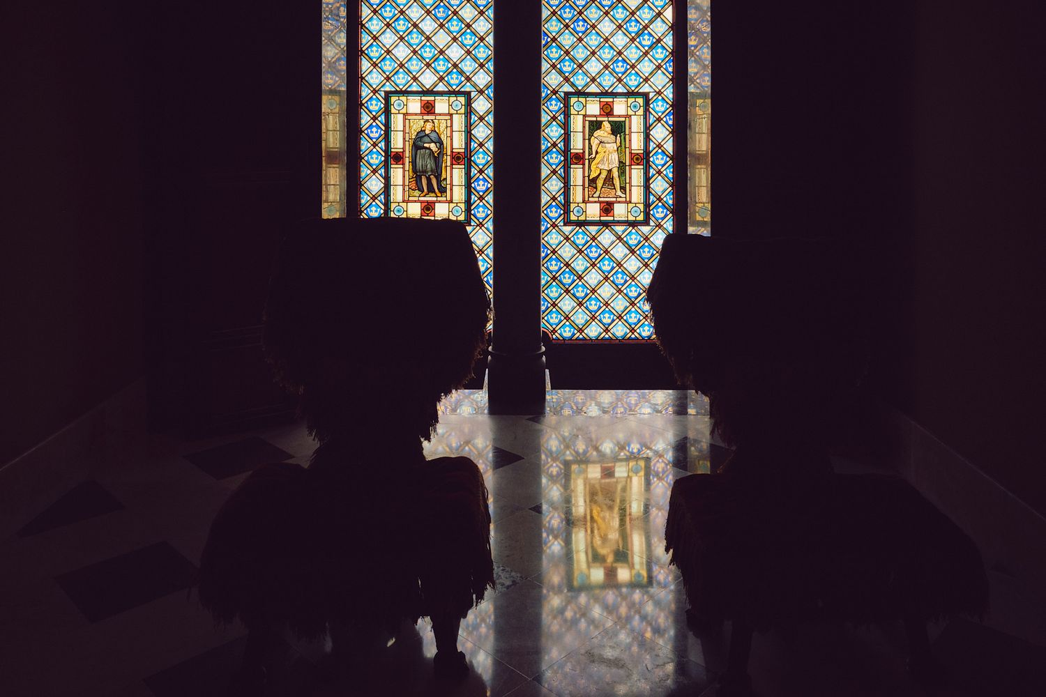 Silhouettes of people in a dark church with colorful stained glass windows depicting saints in background.