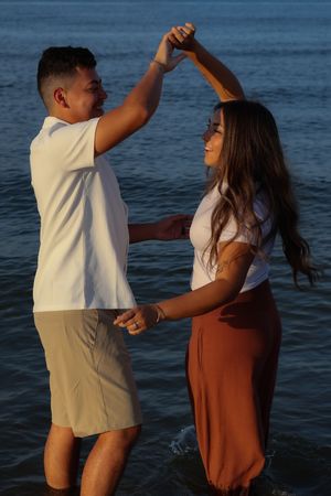 Happy couple dancing together in the ocean water at sunset, wearing white tops and casual summer attire.