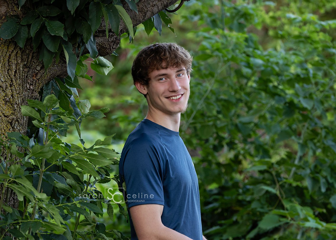 Young man with curly hair smiling outdoors, leaning against a tree surrounded by lush green foliage.