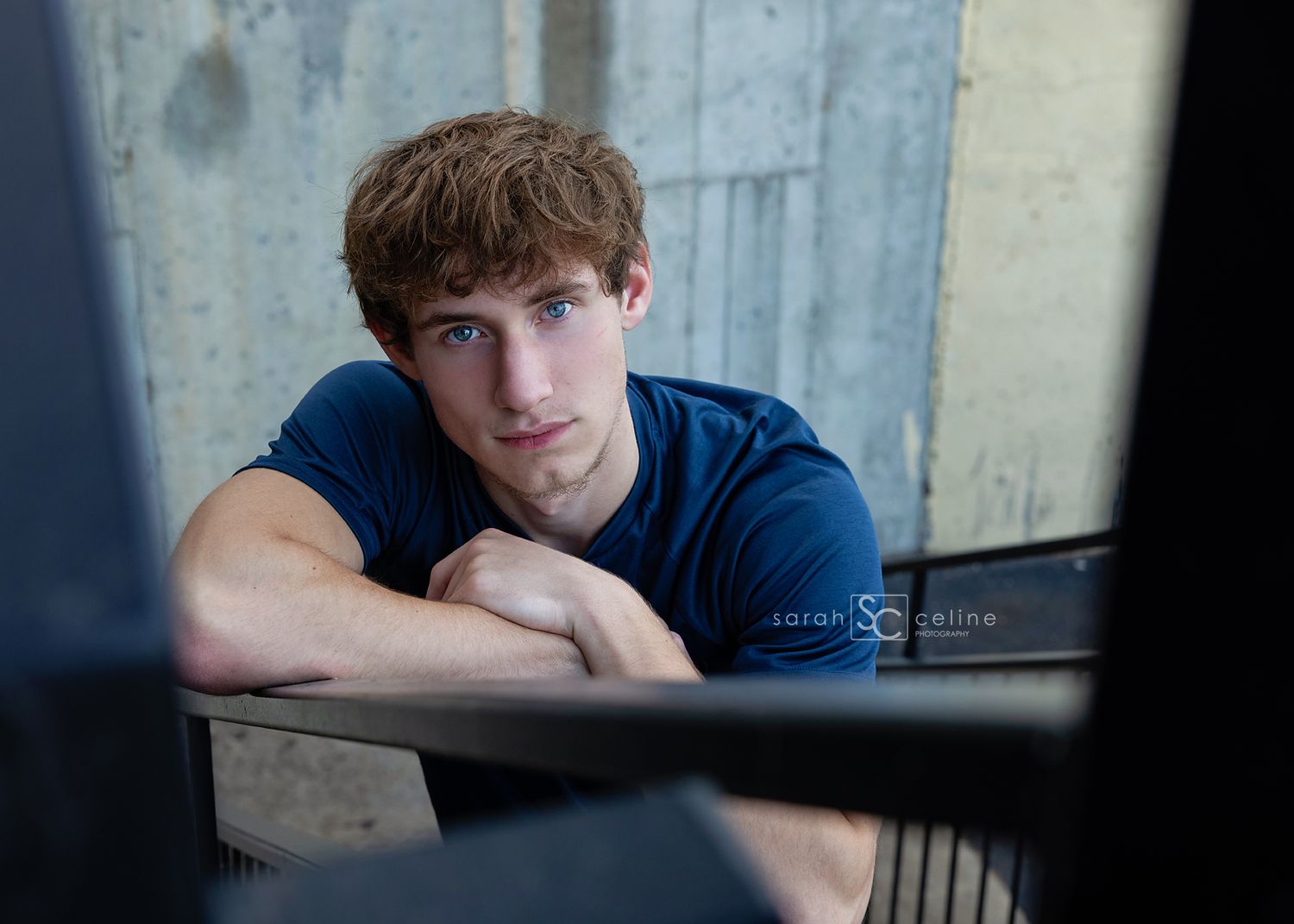 Young man in blue t-shirt leaning on railing, gazing seriously at camera in an urban setting.