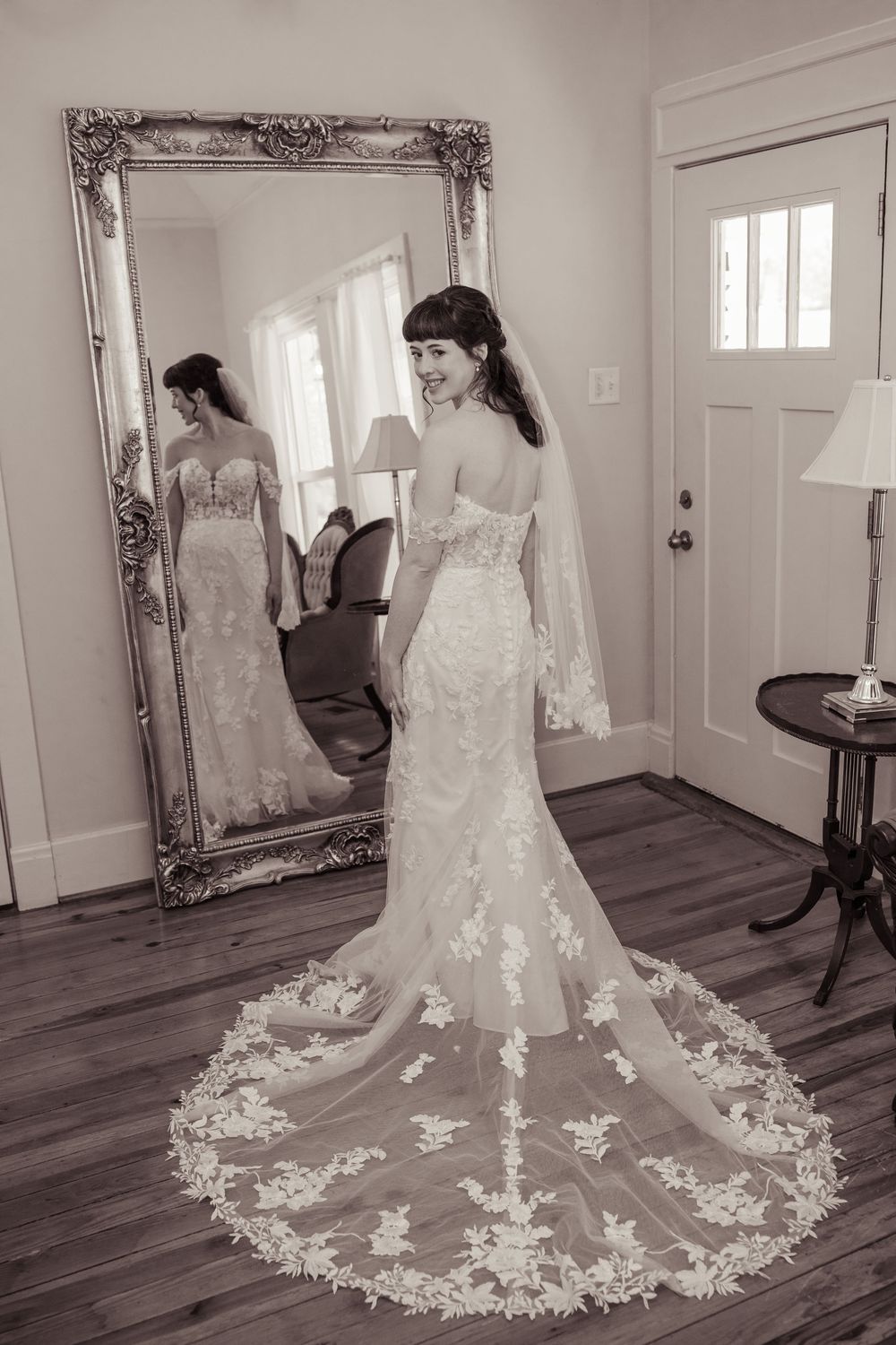 Bride in lace wedding gown with cathedral veil stands before ornate gold mirror in black and white bridal portrait.