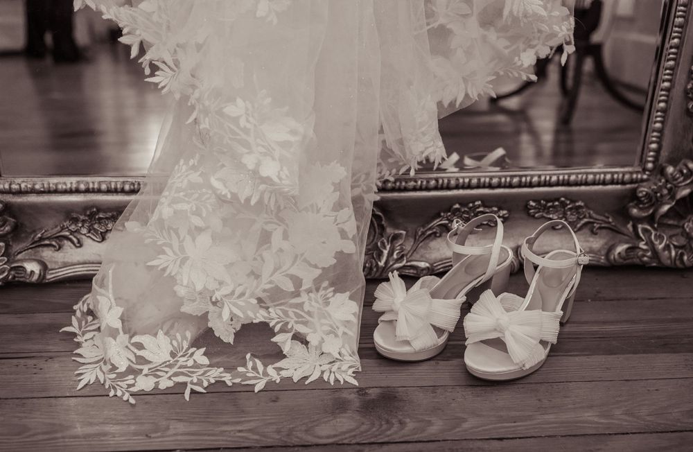 Black and white photo of a lace wedding dress hem and white bridal sandals on a wooden floor near a gilded frame.