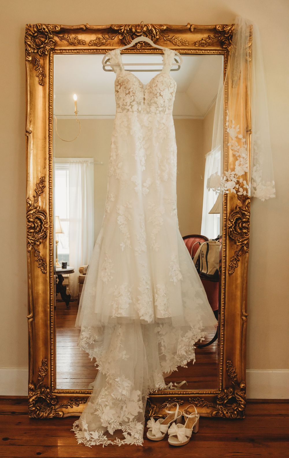 Elegant lace wedding dress hanging on ornate gold mirror with bridal heels displayed below on hardwood floor.