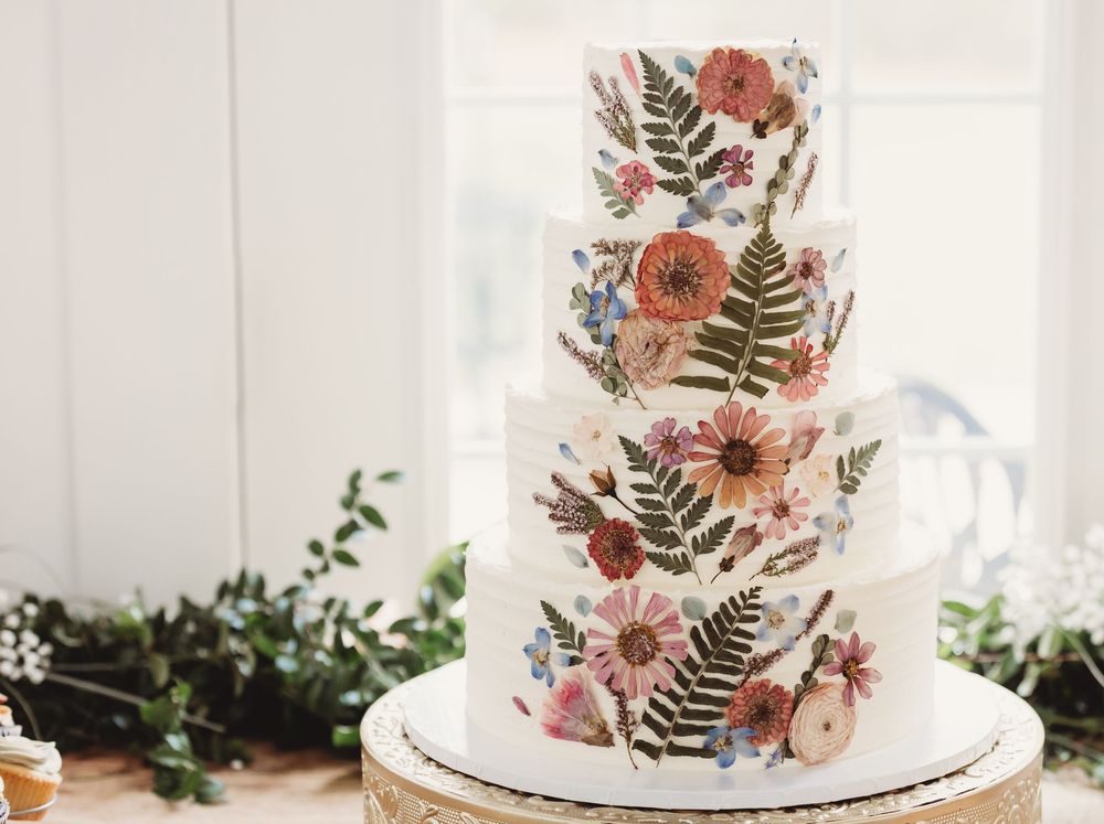 Three-tier white wedding cake adorned with pressed wildflowers and ferns in pink, coral, and blue hues.