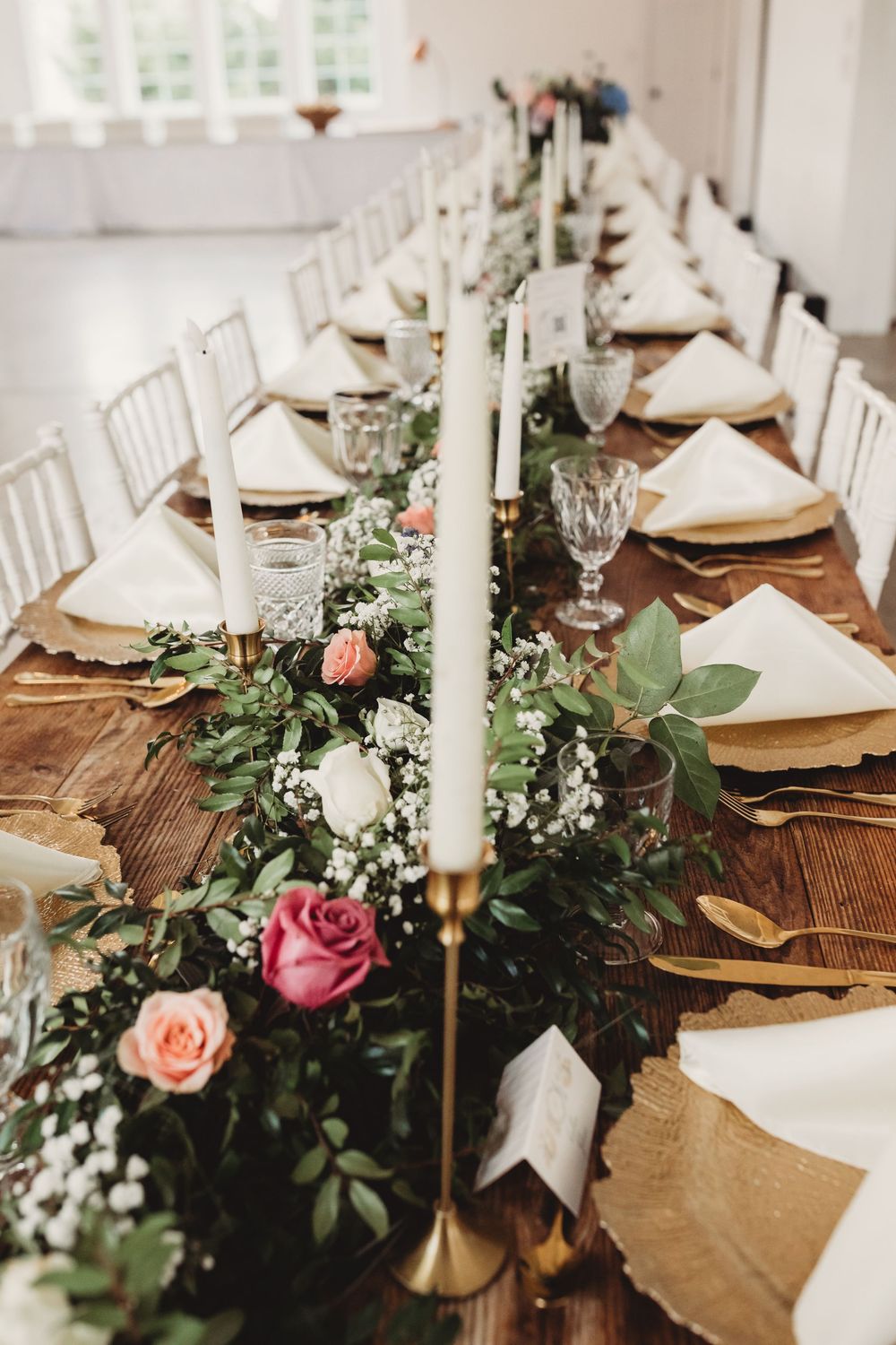 Elegant wedding reception table with floral garland, white taper candles, gold chargers, crystal glasses, and pink roses on rustic wood.