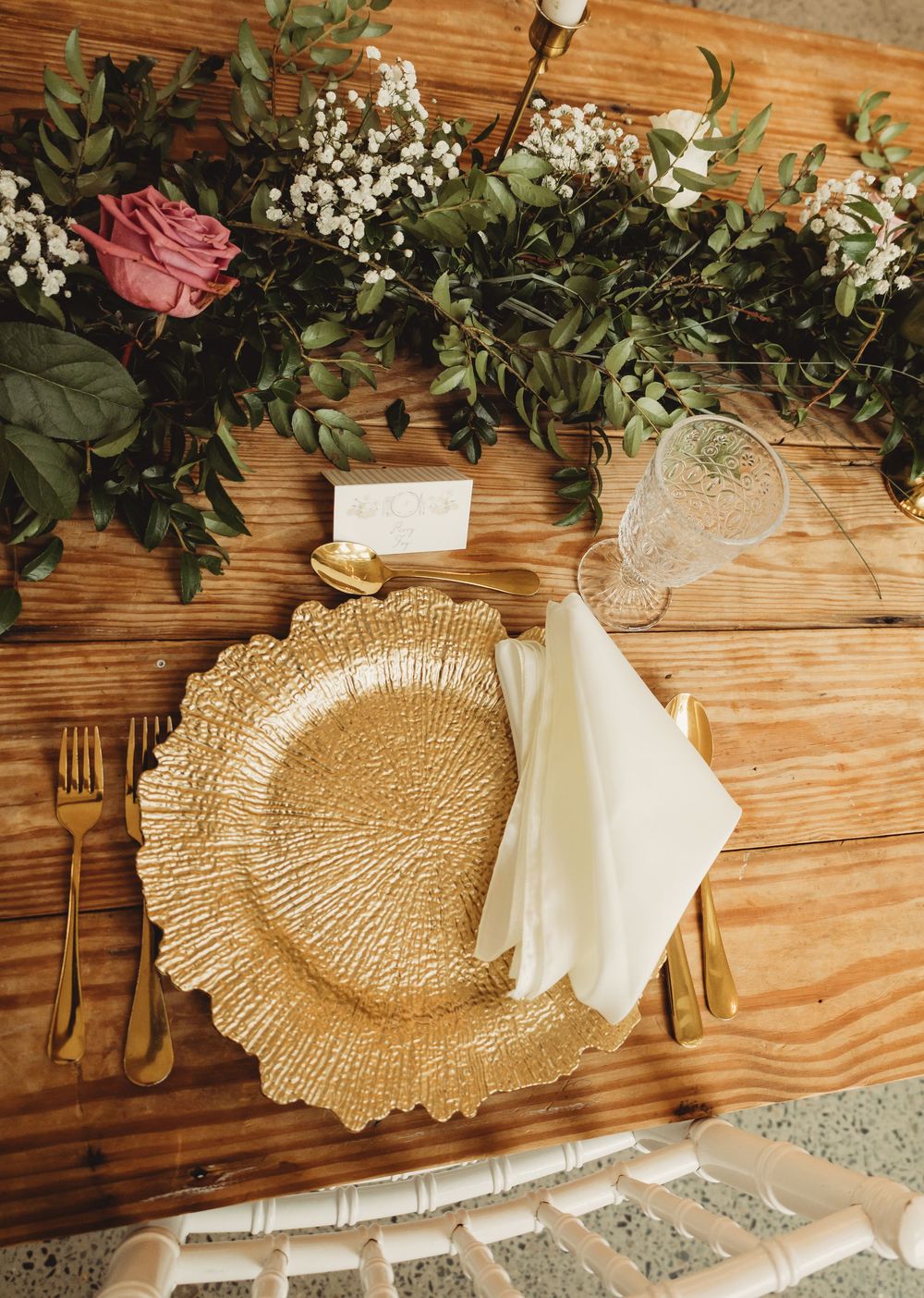 Gold charger plate with white napkin, gold cutlery, crystal glass, and floral greenery centerpiece on rustic wood table.