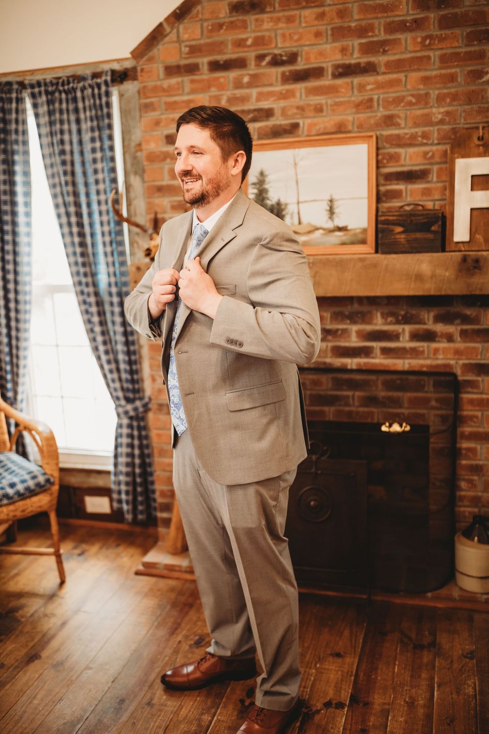 Smiling groom in tan suit adjusting tie in rustic room with brick fireplace and hardwood floors.