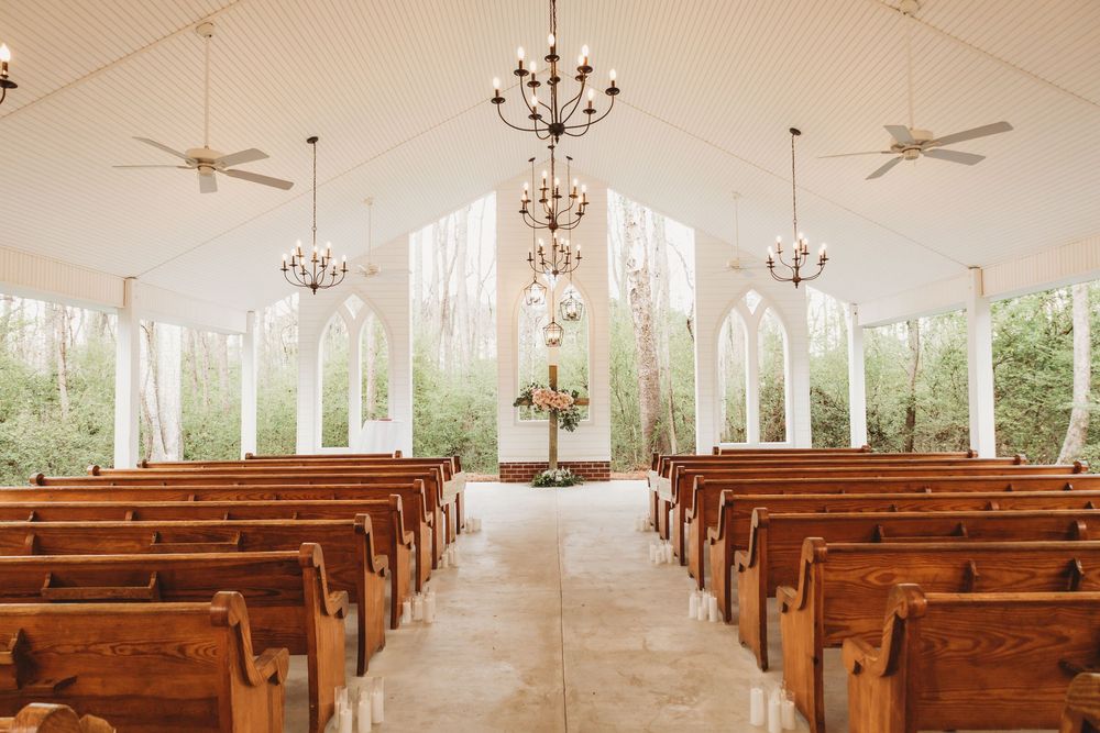 Elegant white chapel wedding venue with wooden pews, arched windows, and chandelier lighting surrounded by trees.