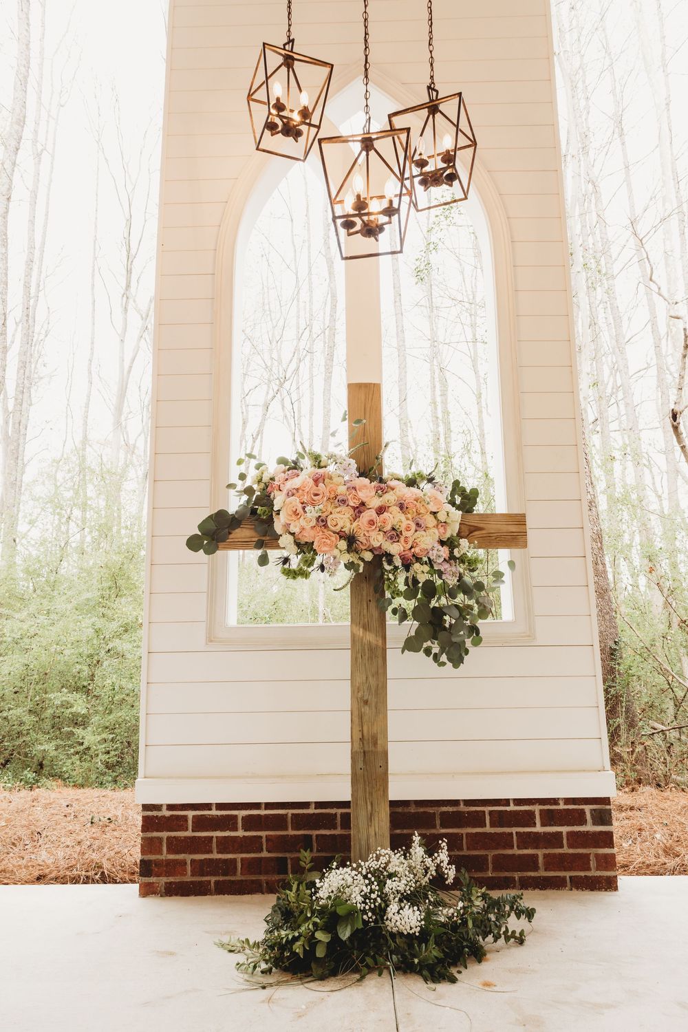 Wooden cross adorned with pink roses and greenery stands before a white chapel with geometric chandeliers in a forest setting.