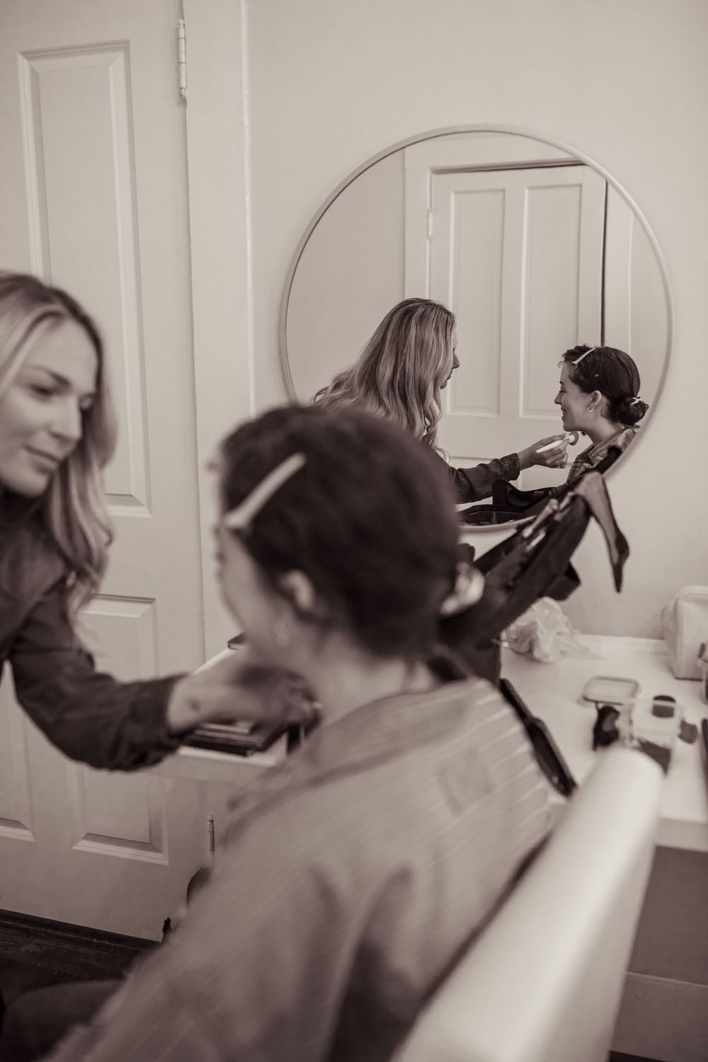 Bride getting hair and makeup done by stylist, reflected in round mirror, sepia-toned bridal preparation photo.