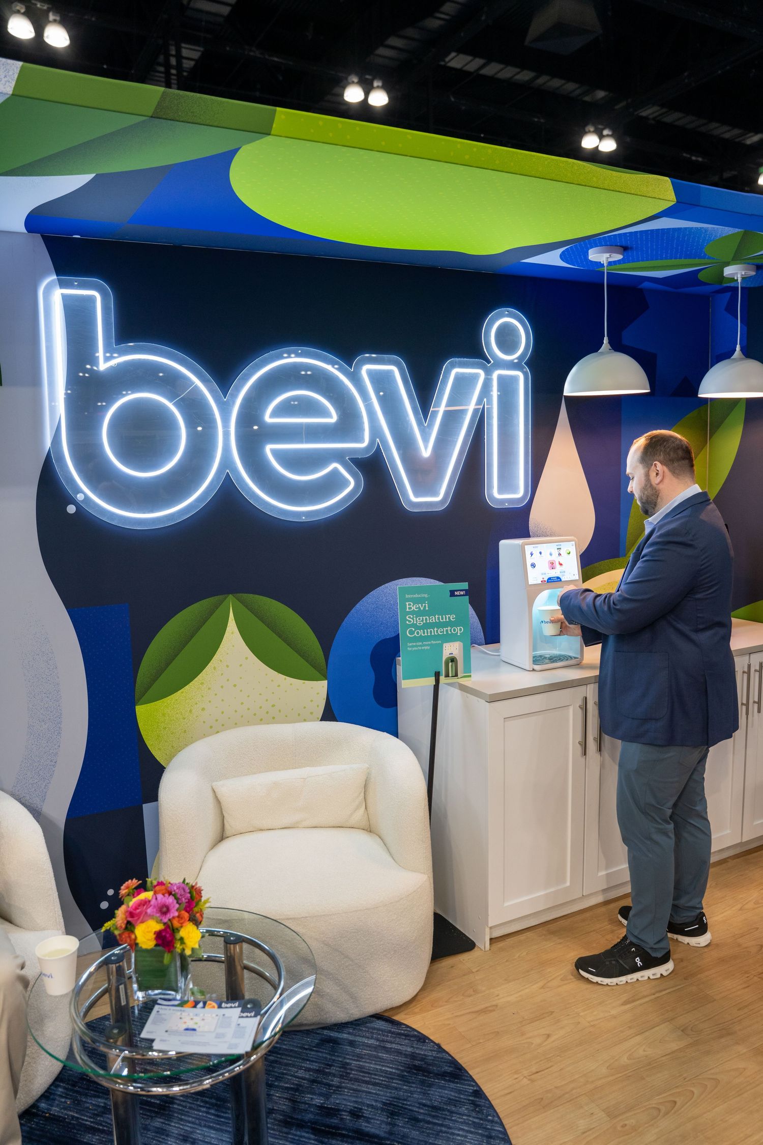 Man using a Bevi smart water dispenser at a colorful trade show booth with neon logo signage.