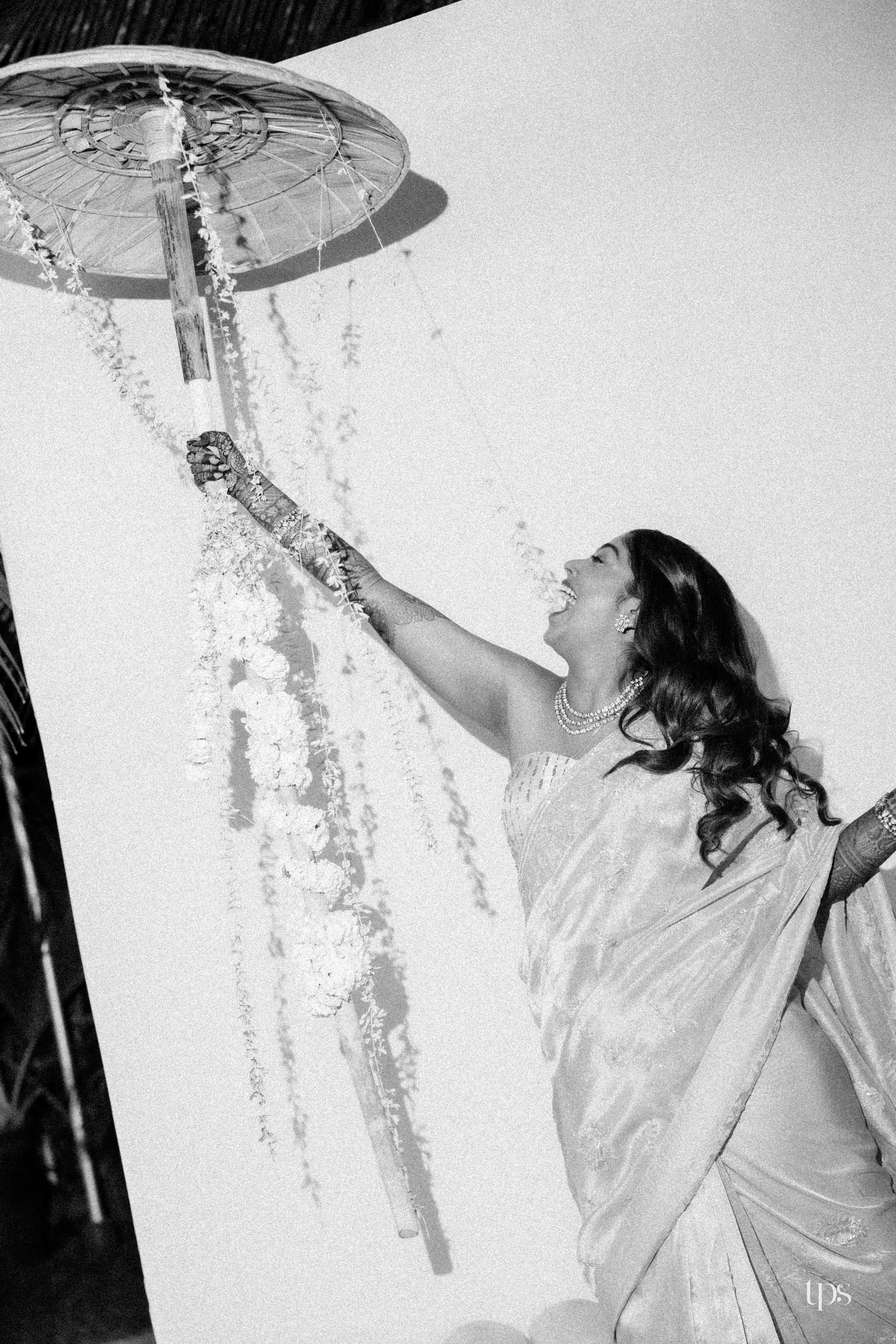 Indian bride in saree joyfully reaching up to a floral chandelier in a black and white photo.