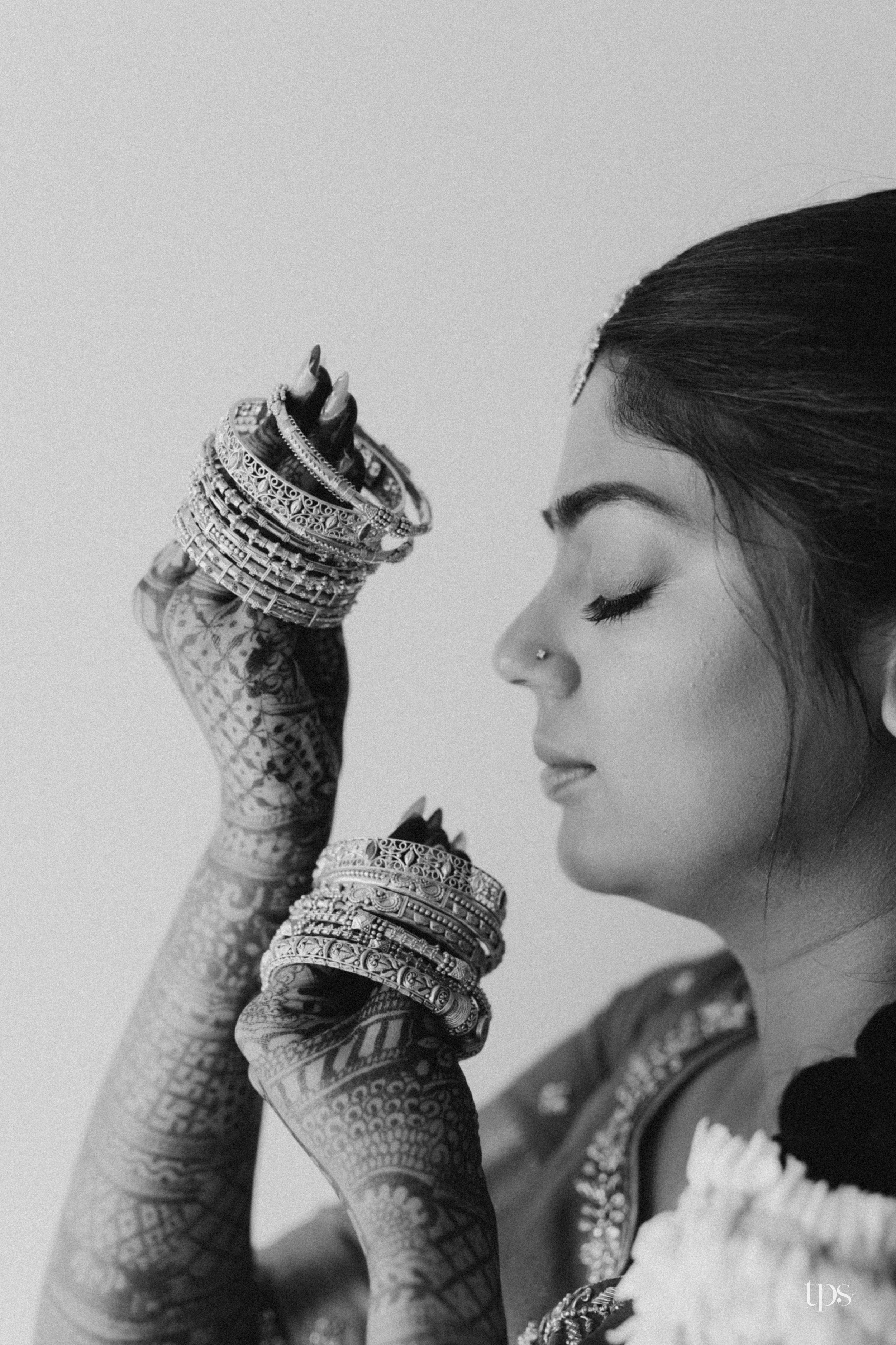 Black and white photo of a bride with henna-covered hands displaying bangles, eyes closed, wearing a nose ring.