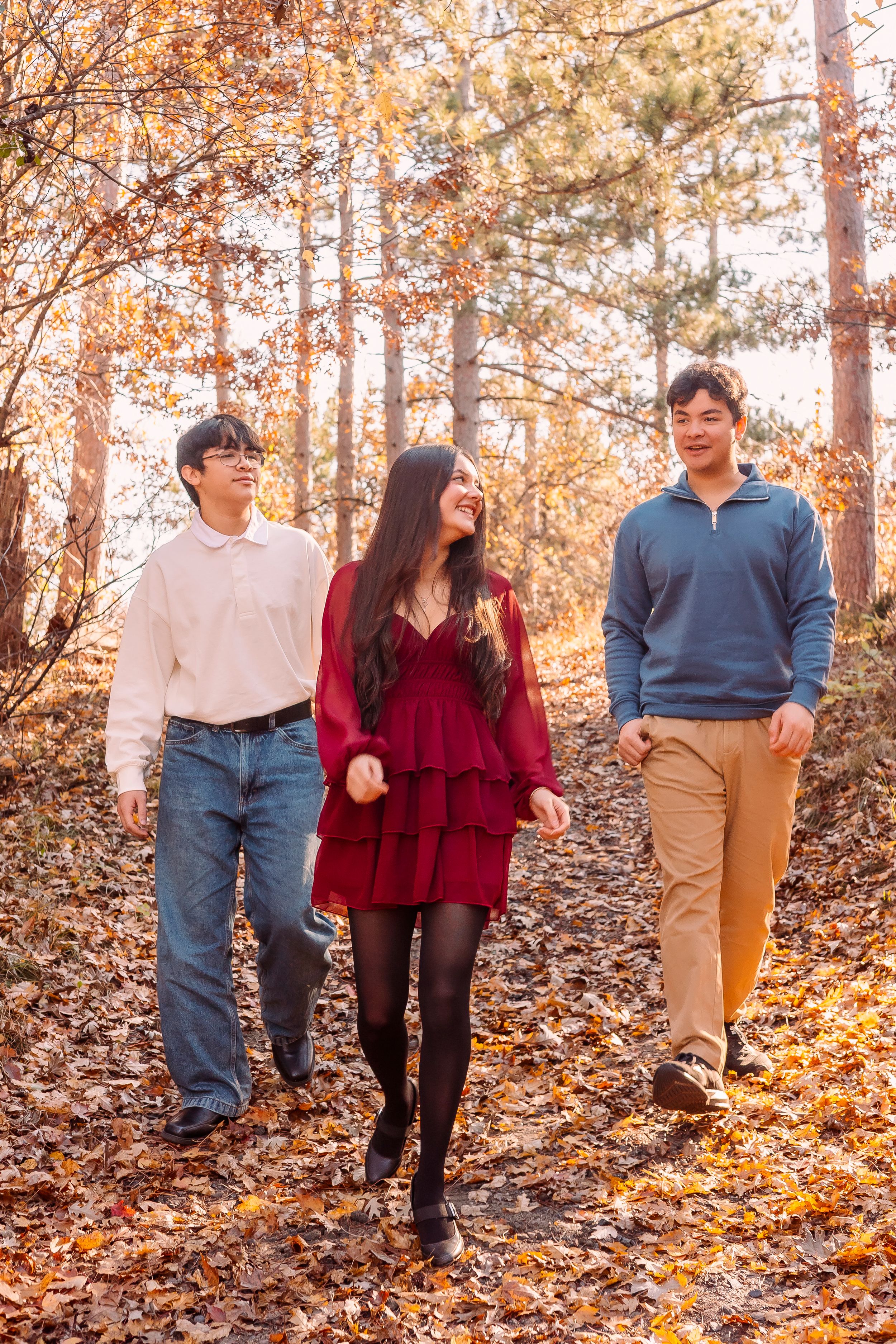 Three young adults walk through an autumn forest path, smiling, wearing casual fall outfits among colorful fallen leaves.