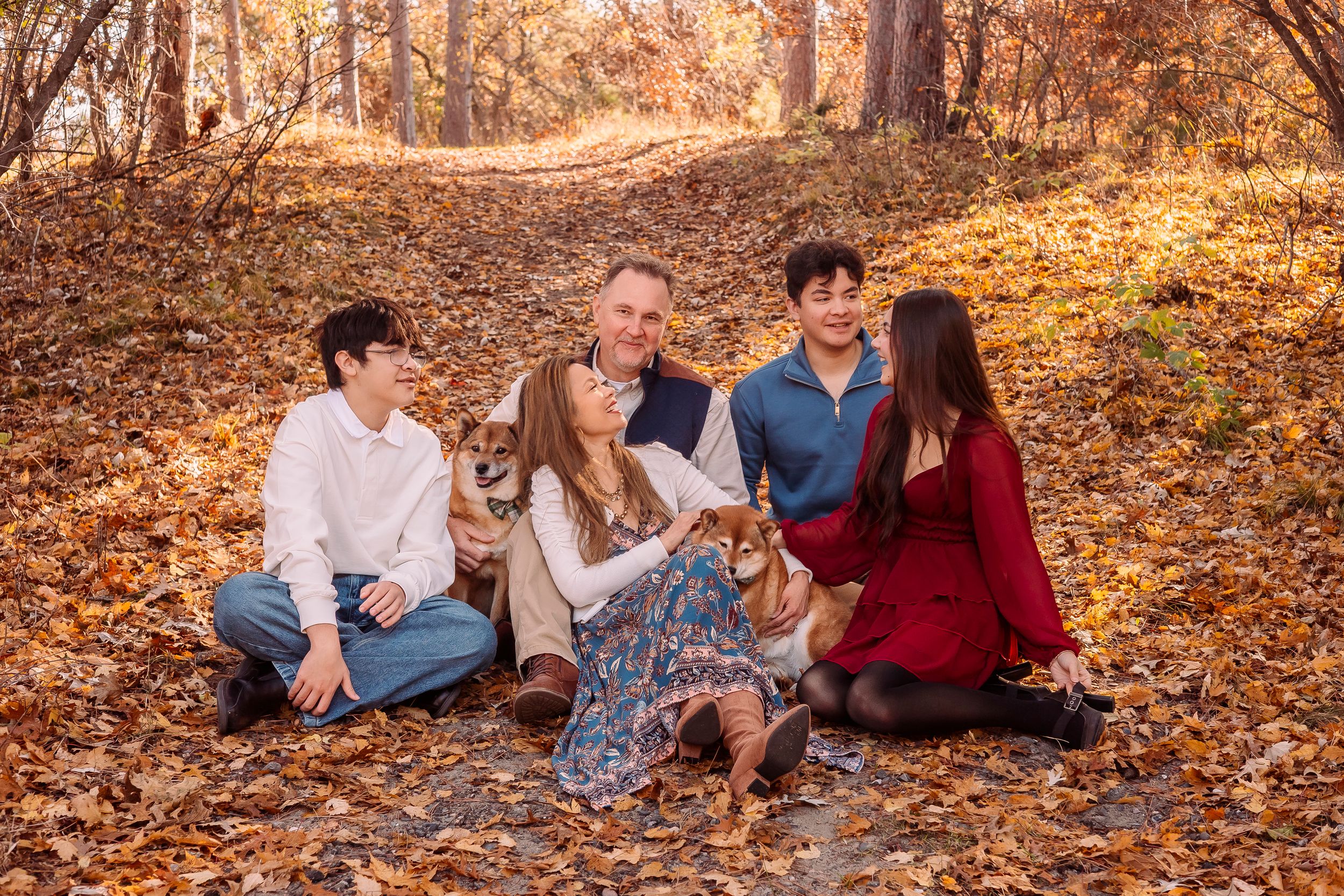 Family of five sitting among autumn leaves in a forest, smiling and enjoying fall foliage together.