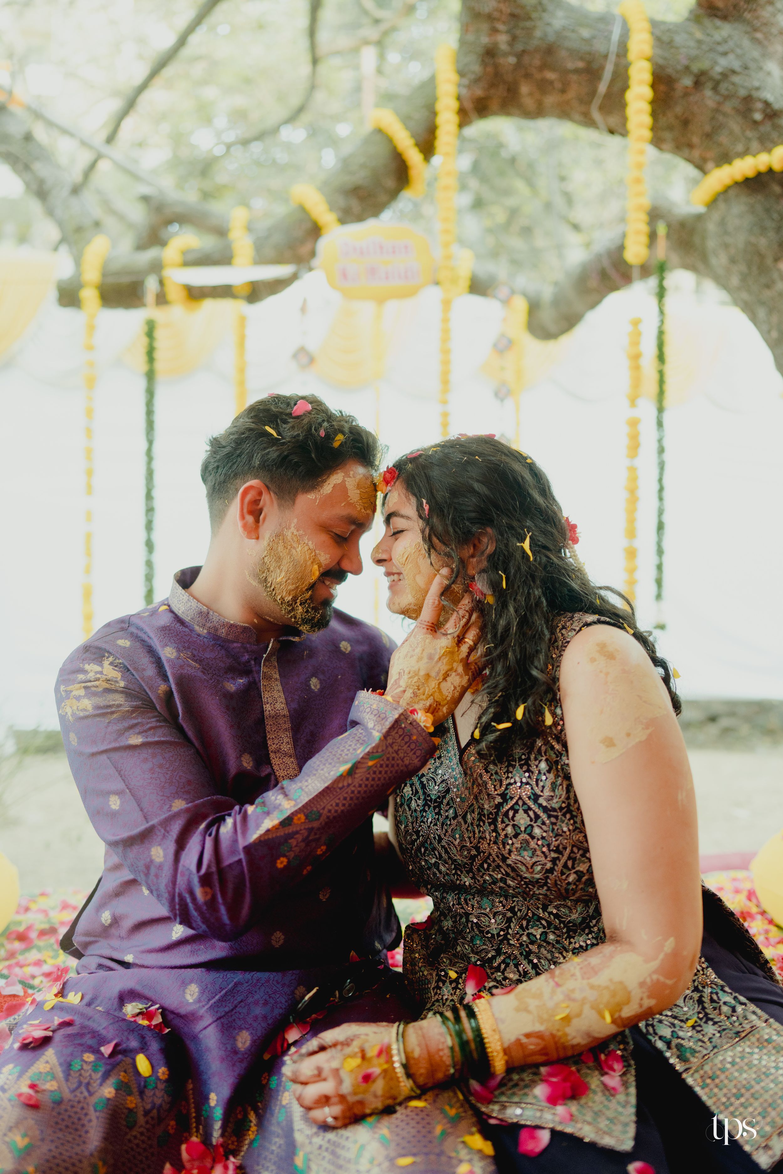 Indian couple celebrating haldi ceremony under decorated tree, covered in colorful flower petals and turmeric.