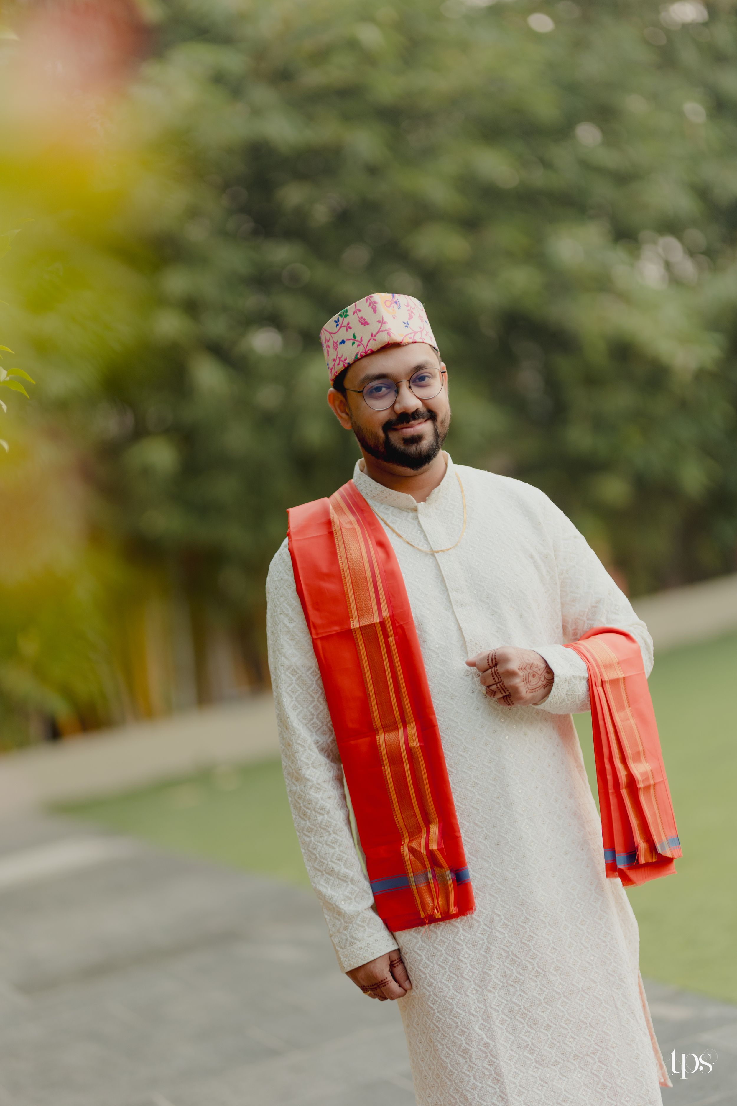 Man in white kurta, red dupatta, and embroidered topi smiling outdoors at a traditional Indian wedding.
