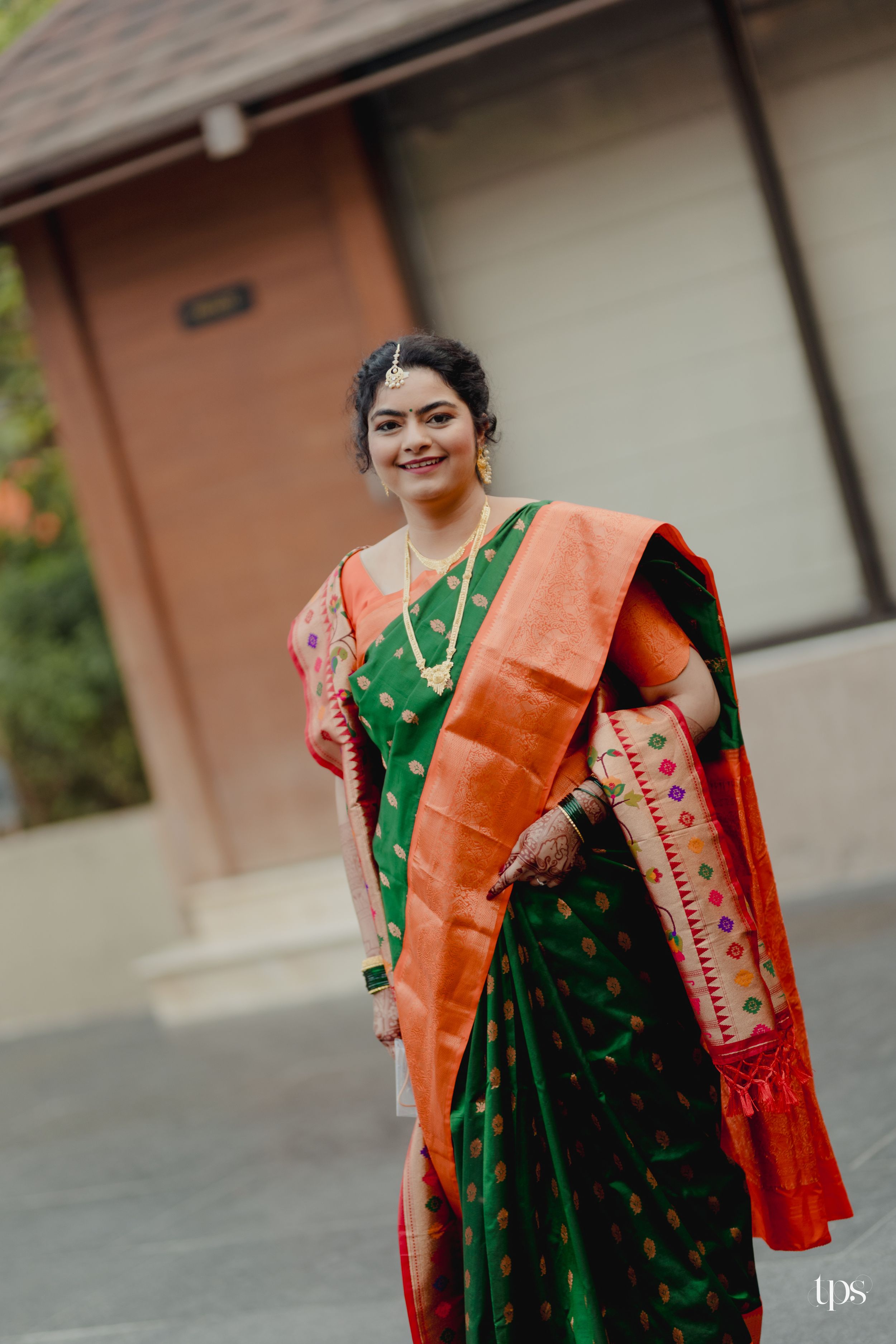 Bride in green and orange Paithani silk saree with gold jewelry smiling outdoors.