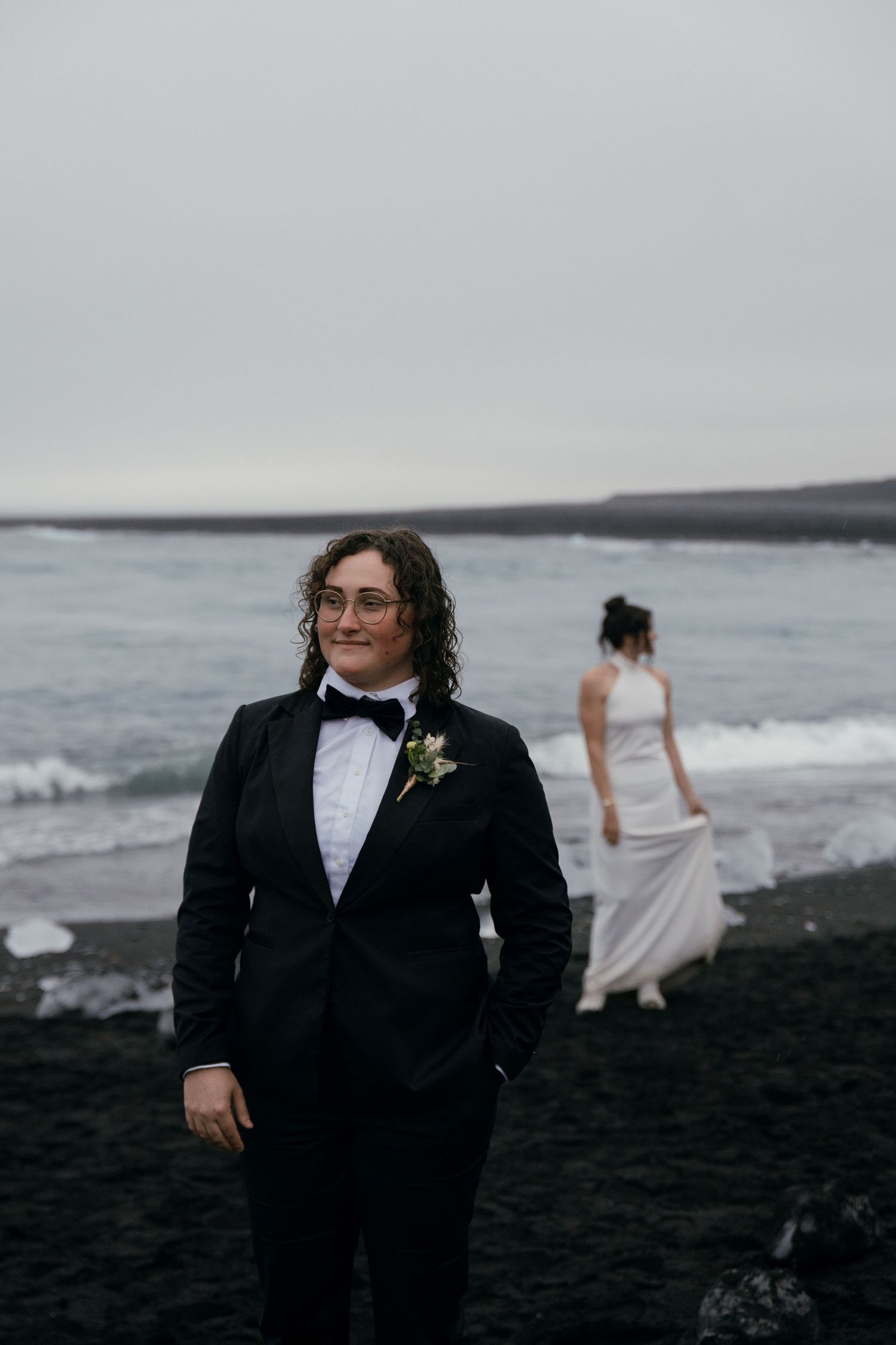 Two brides on black sand beach, one in tuxedo with boutonniere, other in white dress, ocean backdrop.