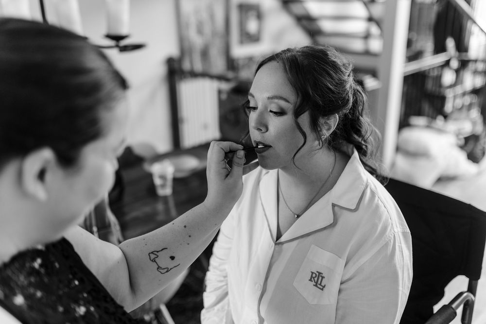 Black and white photo of a bride getting lipstick applied by a makeup artist while wearing a monogrammed robe.