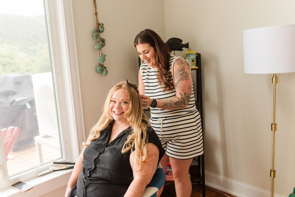 Woman getting hair styled by tattooed stylist in striped dress near bright window in modern home.