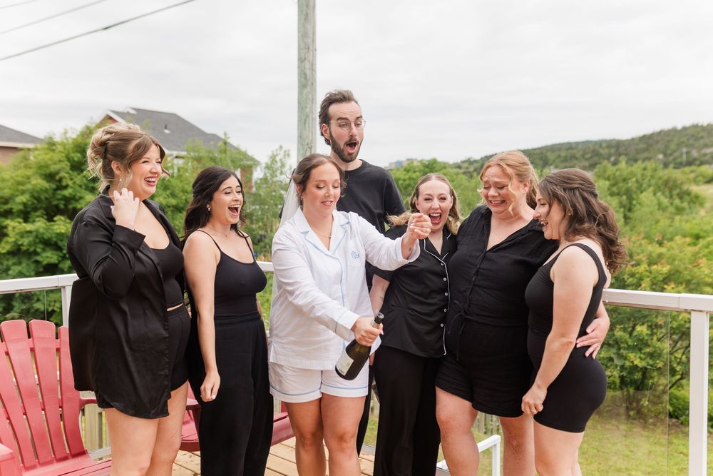 Bride in white robe celebrates with bridesmaids in black on a deck, laughing and opening champagne before the wedding.
