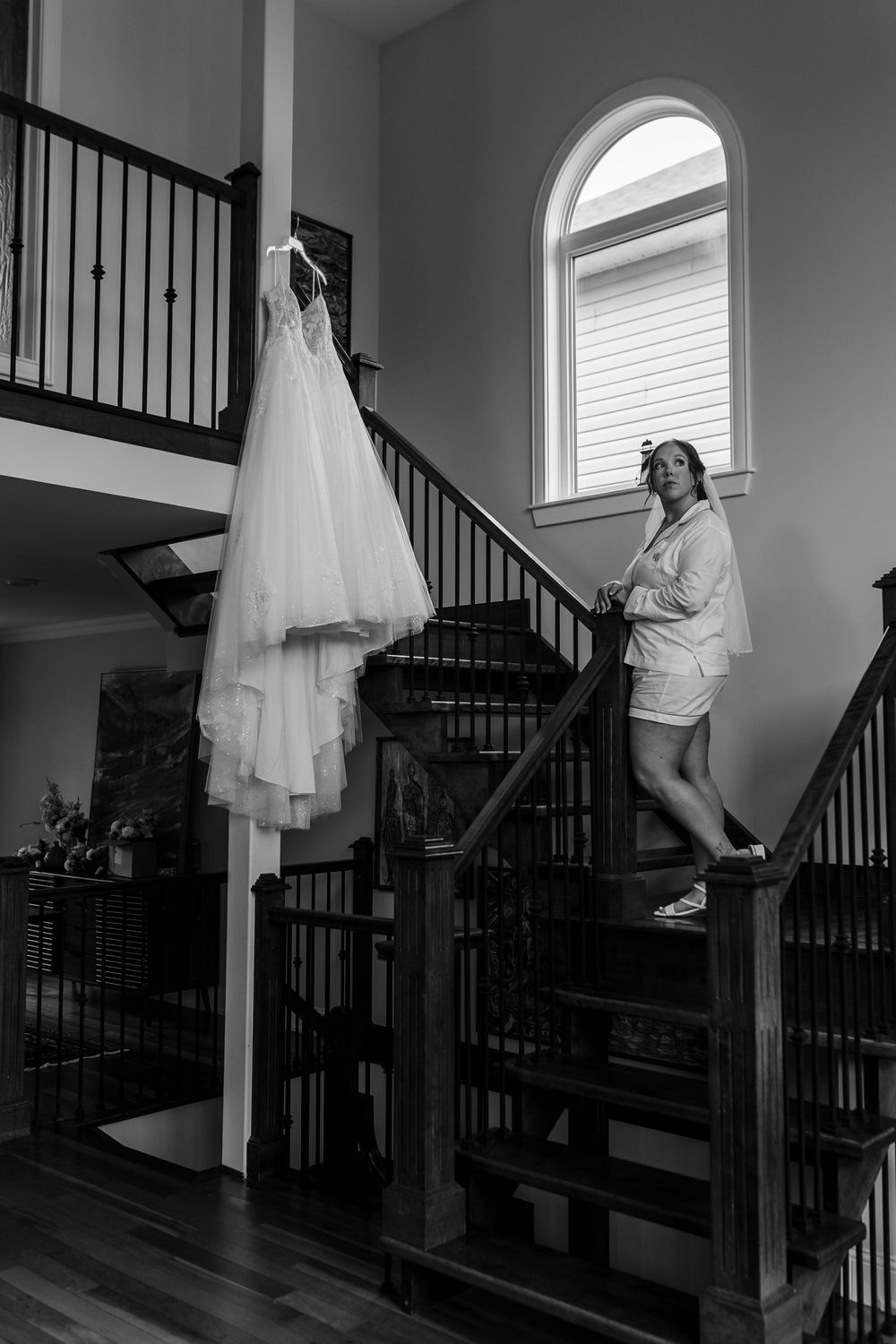 Bride in robe stands on wooden staircase beside hanging wedding gown in elegant home, black and white photo.