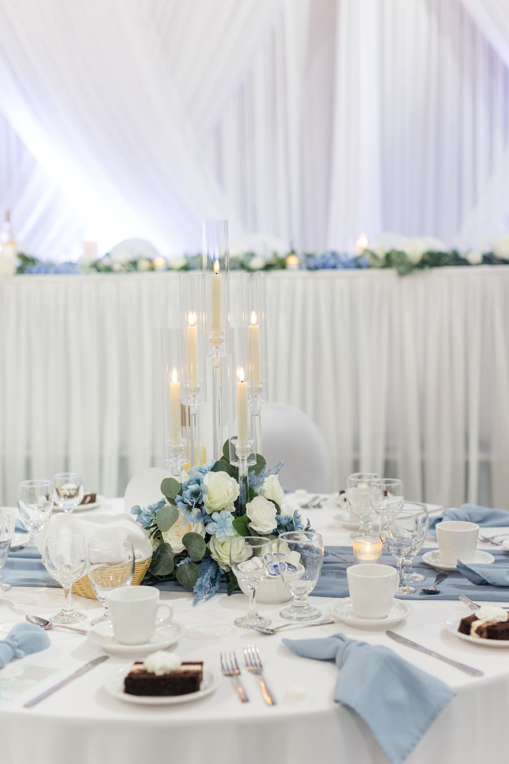 Elegant wedding reception table with blue napkins, white roses, candelabra centerpiece, and dessert plates in a draped venue.