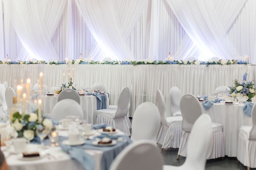 Elegant wedding reception hall with white draping, blue accents, floral centerpieces, and candlelit round tables.