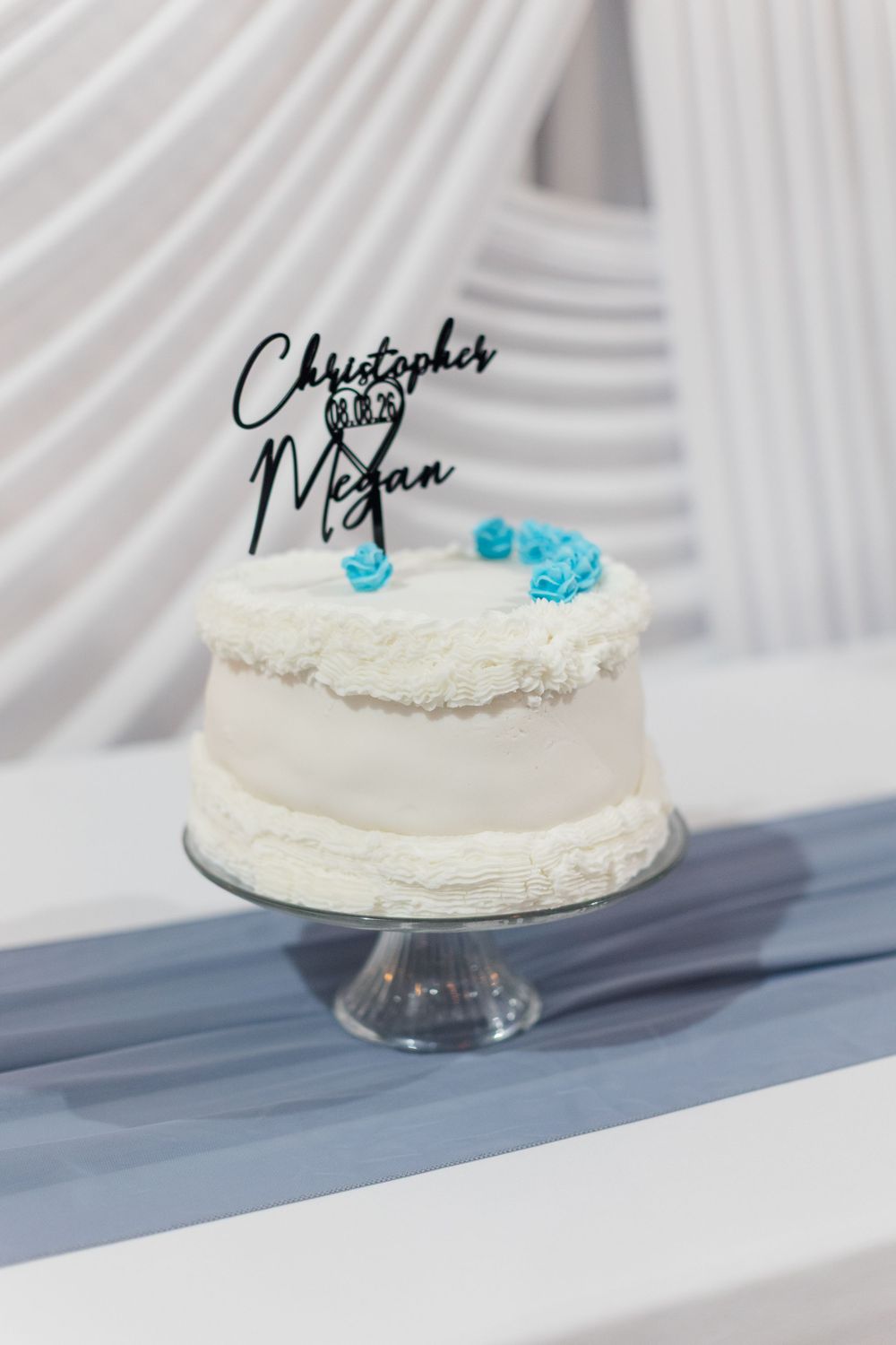White wedding cake with blue roses and Christopher & Megan cake topper on a glass stand.