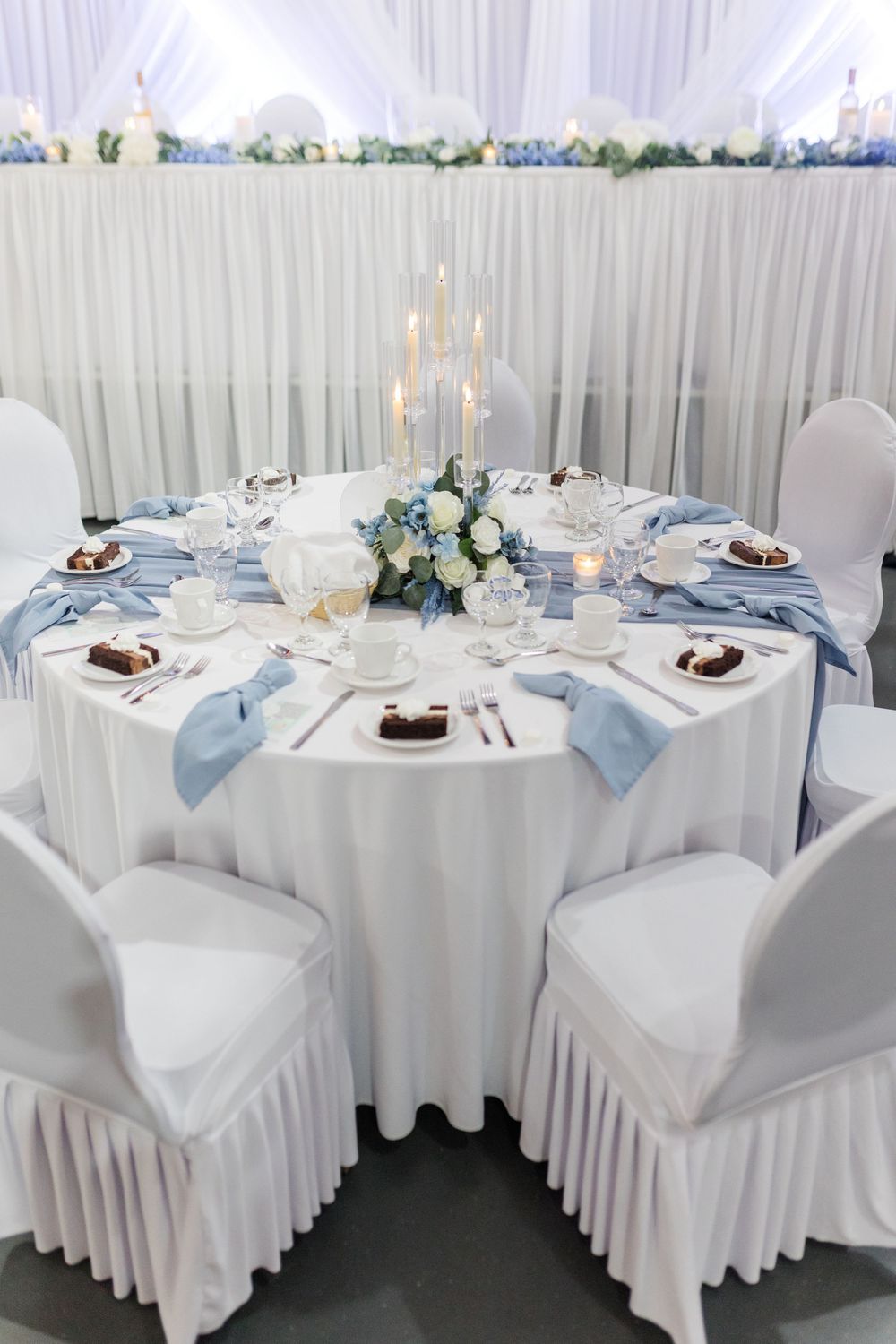 Elegant wedding reception table with white linens, blue satin runner, floral centerpiece, candelabra, and place settings.