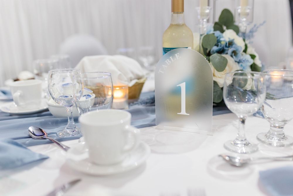 Elegant wedding reception table setting with blue accents, wine glasses, white flowers, candles, and a number one centerpiece.