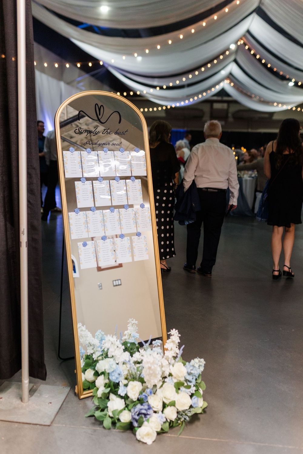 Gold arched wedding seating chart display with white and blue floral arrangement at base, string lights and draped ceiling in background.
