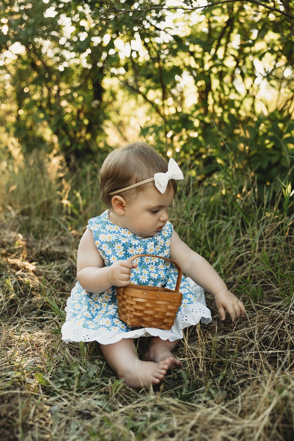Adorable baby girl in blue floral dress with white bow holding wicker basket outdoors.