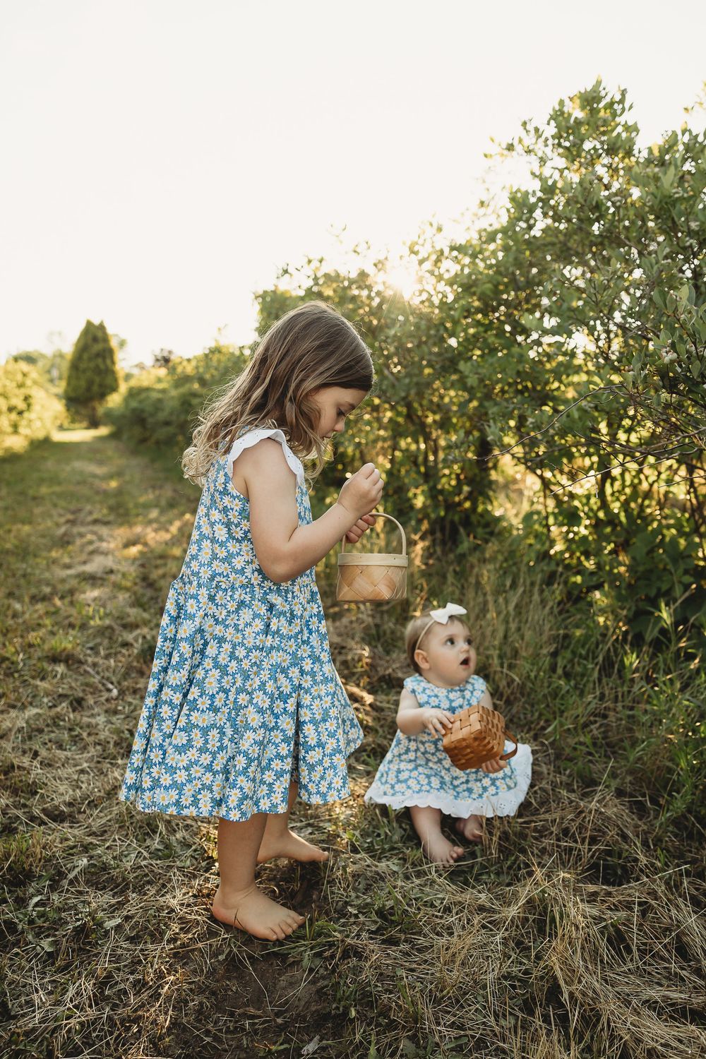 Two barefoot girls in matching blue floral dresses picking berries outdoors at golden hour.