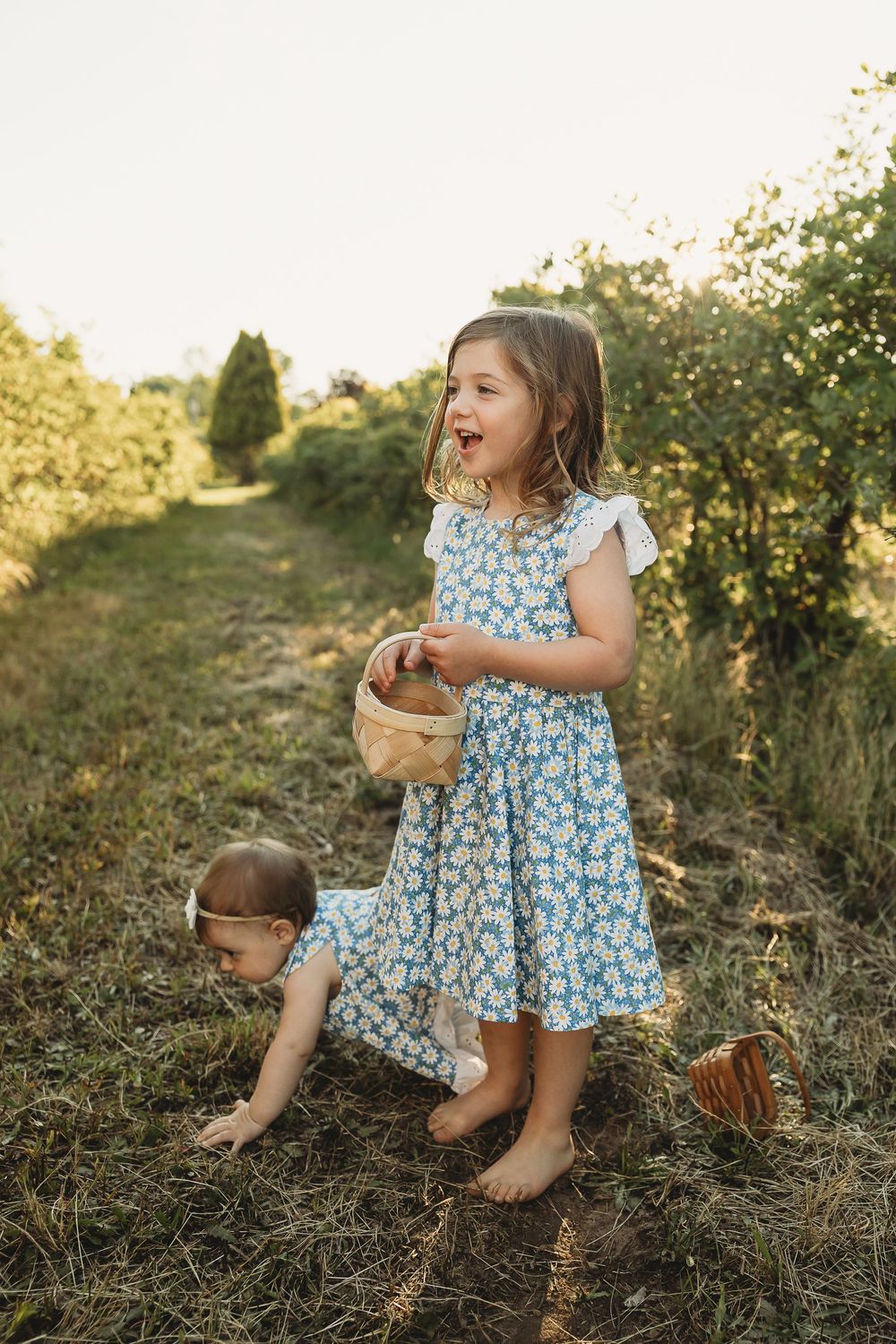 Two young girls in floral dresses picking berries in a sunny garden path, one holding a small wooden basket.