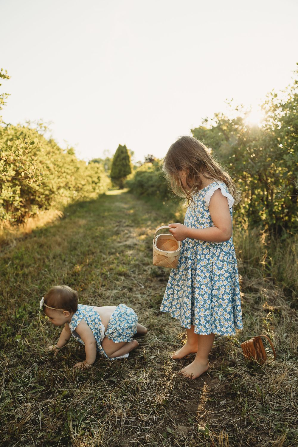 Two young girls in matching floral dresses picking berries in a sunny orchard, one holding a wooden basket.