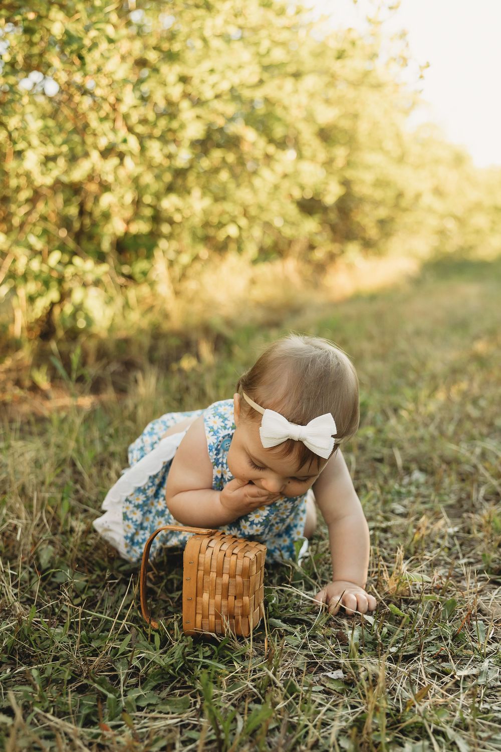 Adorable baby girl in floral dress with white bow crawls on grass holding a wicker basket outdoors.