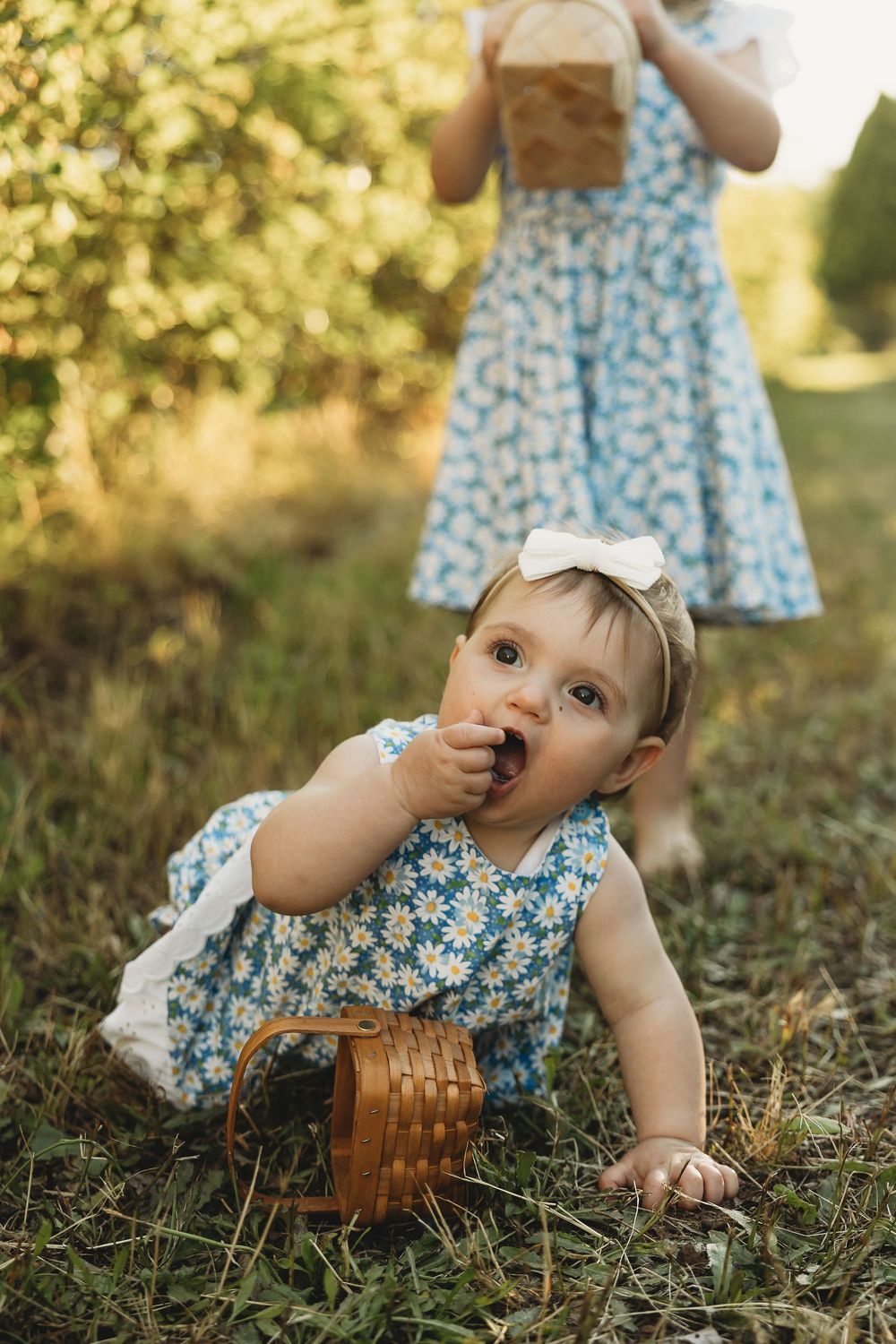 Two young girls in matching blue floral dresses picking berries outdoors, baby sitting on grass eating fruit from a wicker basket.