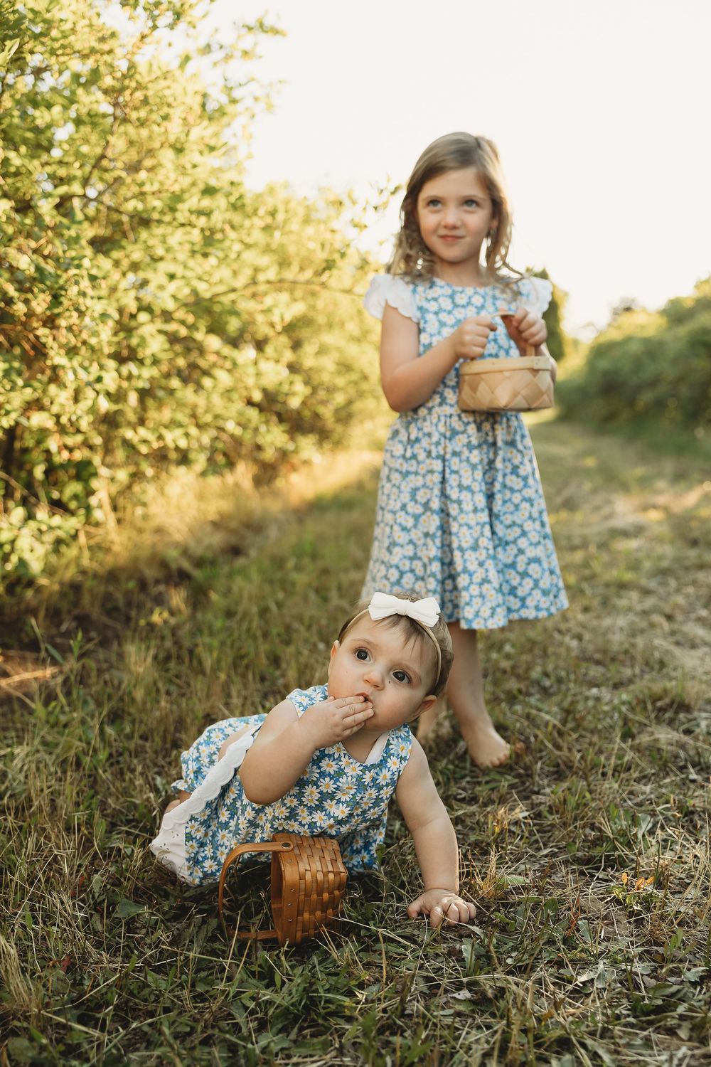 Two young sisters in matching blue floral dresses picking berries outdoors in a sunny garden path.