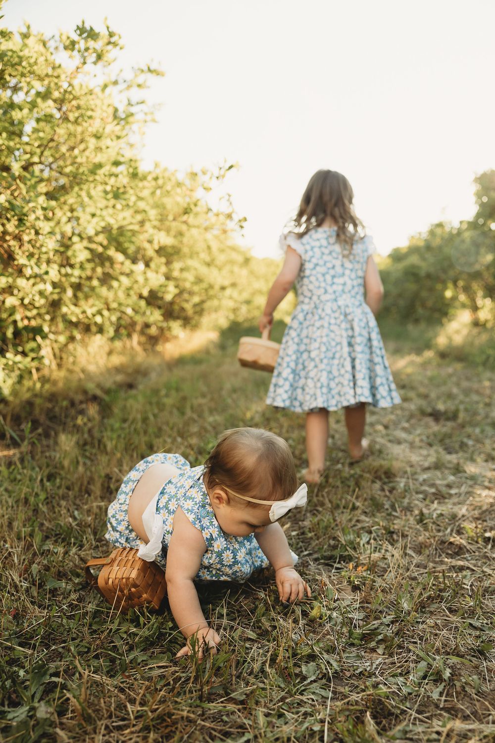 Two young girls in floral dresses picking fruit in a sunny orchard, one crawling on grass with a basket nearby.