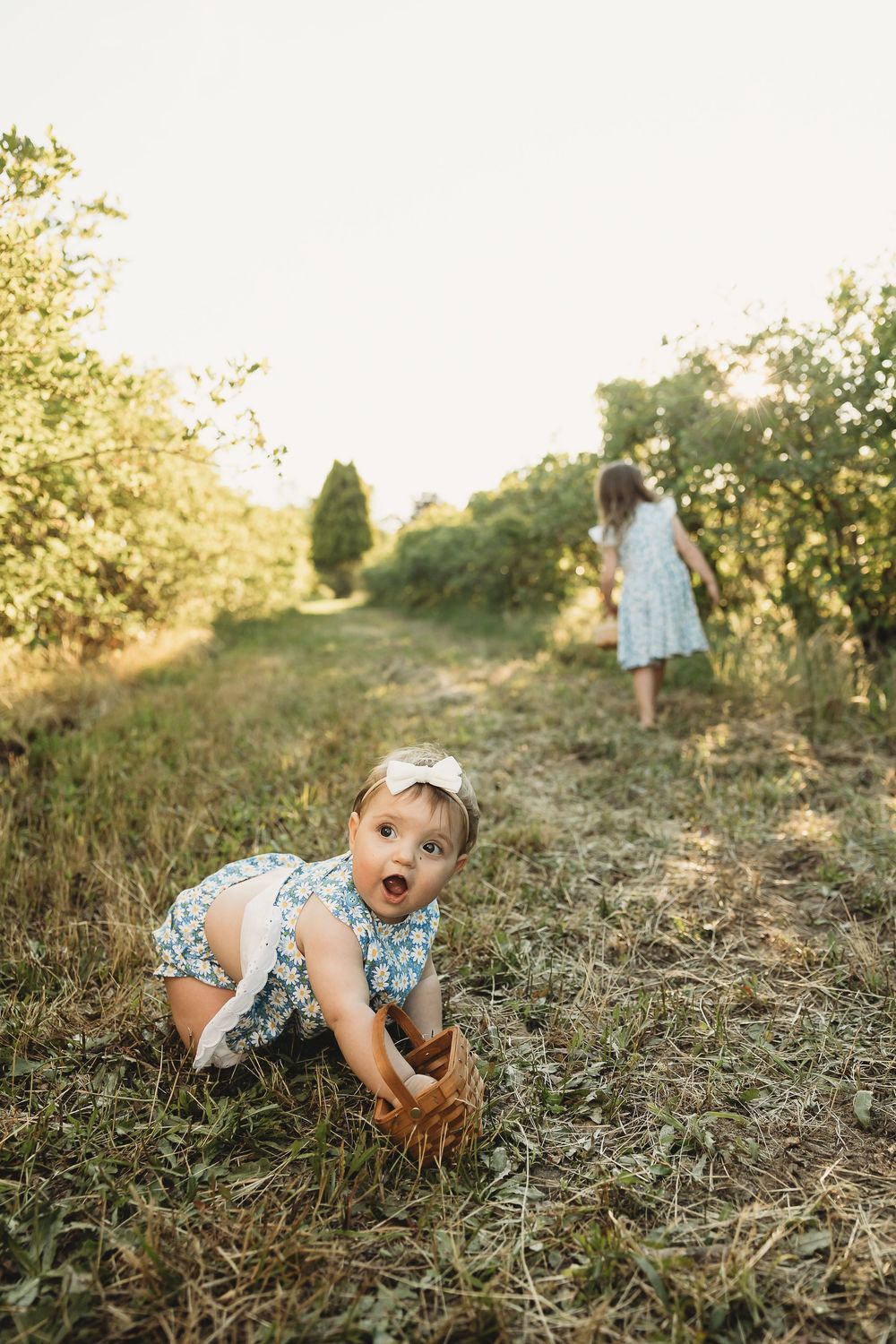 Two young girls in floral dresses playing outdoors in a sunny green orchard, toddler crawling on grass.