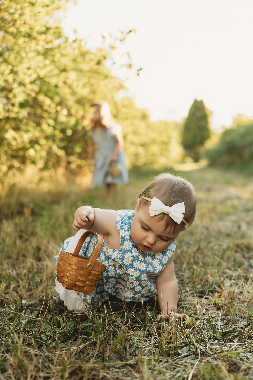 Toddler girl in floral dress with white bow crawls on grass holding wicker basket outdoors in sunny garden.